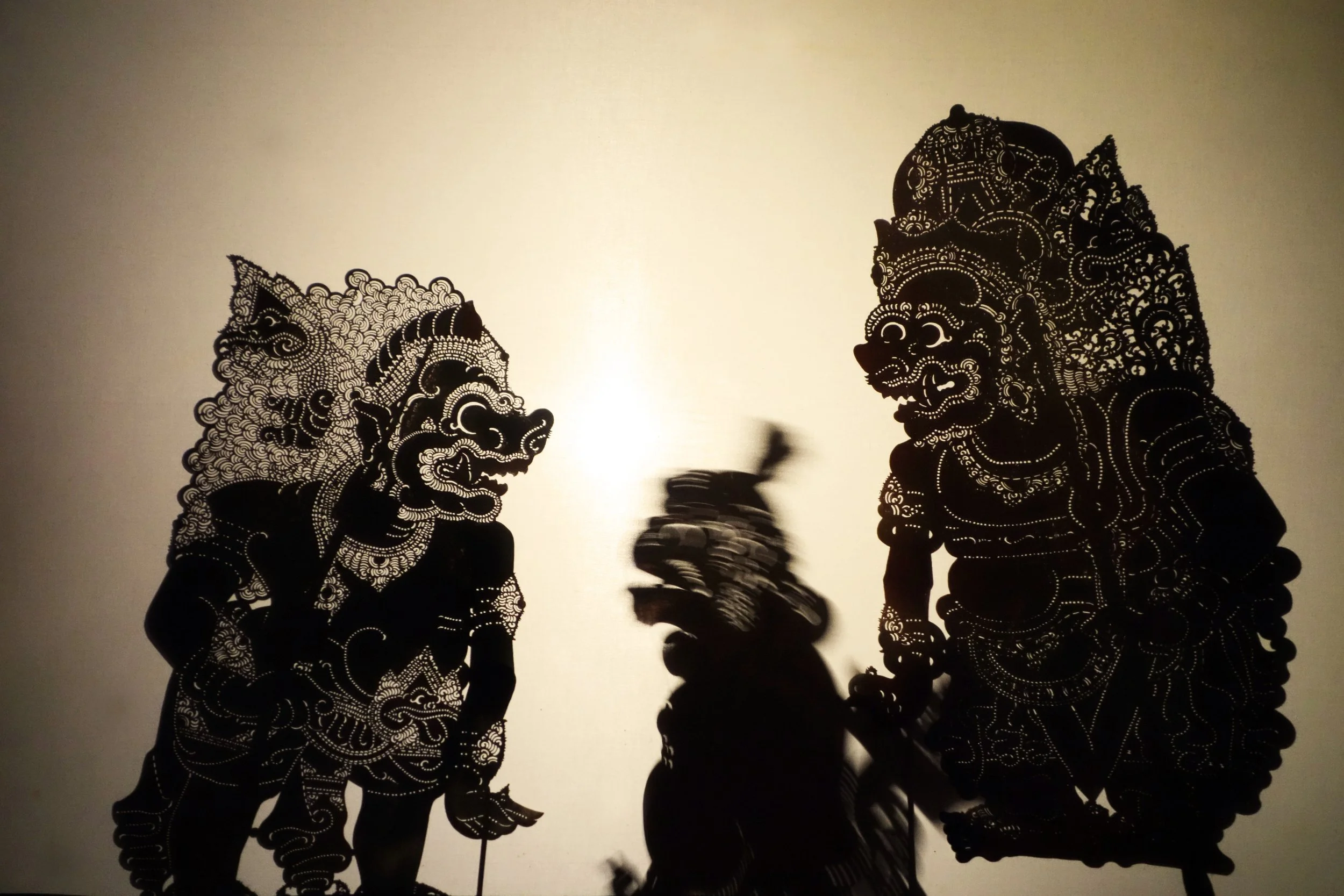 Silhouettes of two traditional Asian mythological figures with intricate details, and a blurred smaller figure between them, all cast as shadows on a light-colored background.