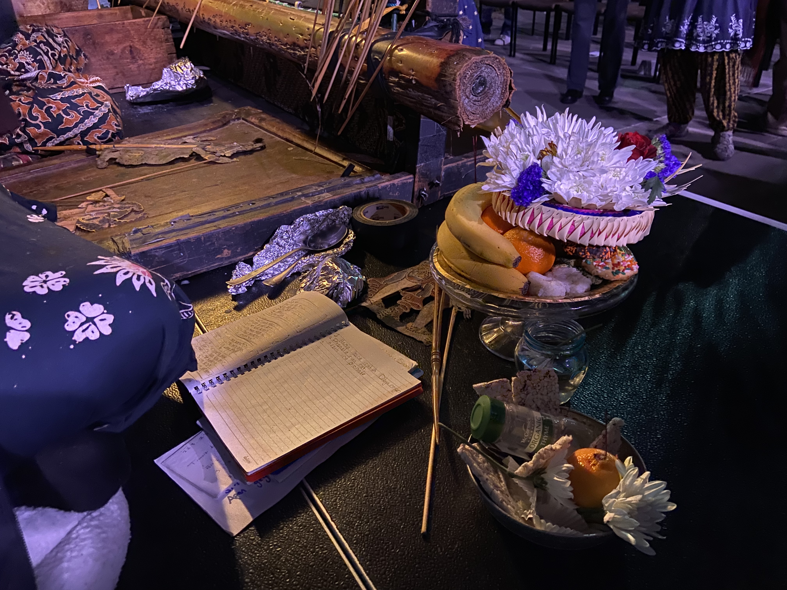 An altar with flowers, fruit, a notebook, and ceremonial items, set on a table with a dark tablecloth at a cultural event.