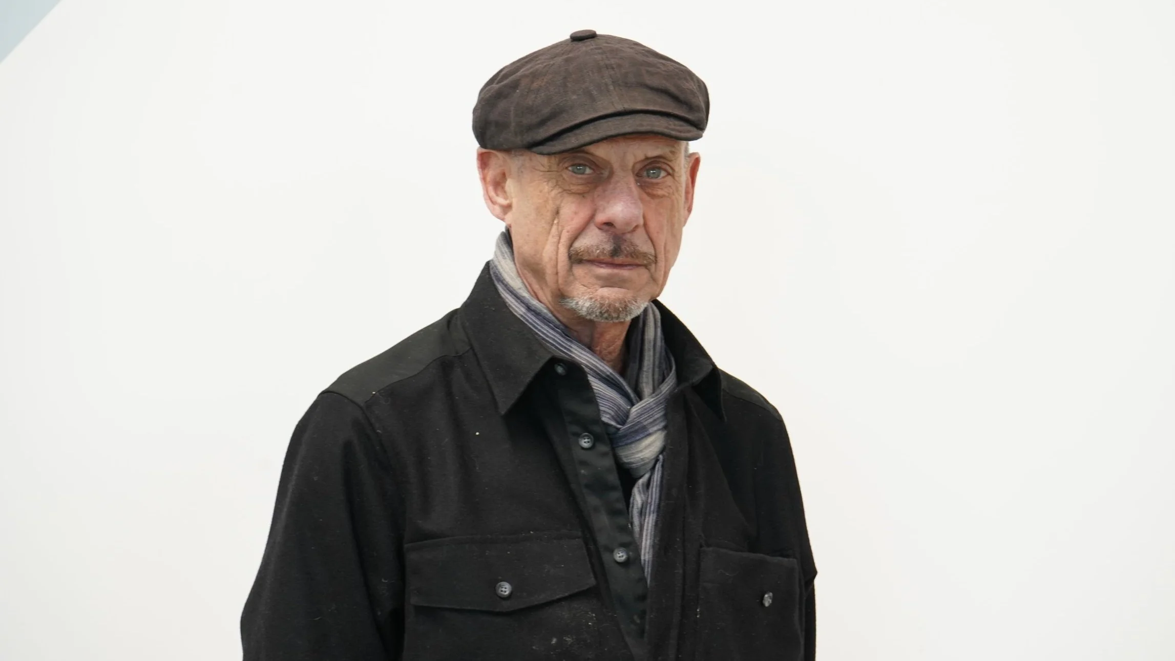An elderly man with gray hair and a goatee, wearing a black button-up shirt, a gray scarf, and a brown flat cap, standing against a plain white background.