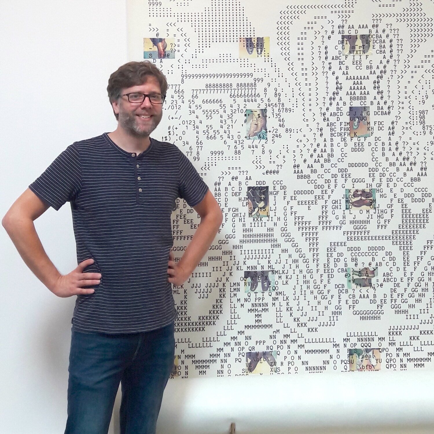 A man with glasses and a beard stands with his hand on his hip in front of a large ASCII art poster of a black panther. The poster contains small images of animals and maps integrated with ASCII characters.