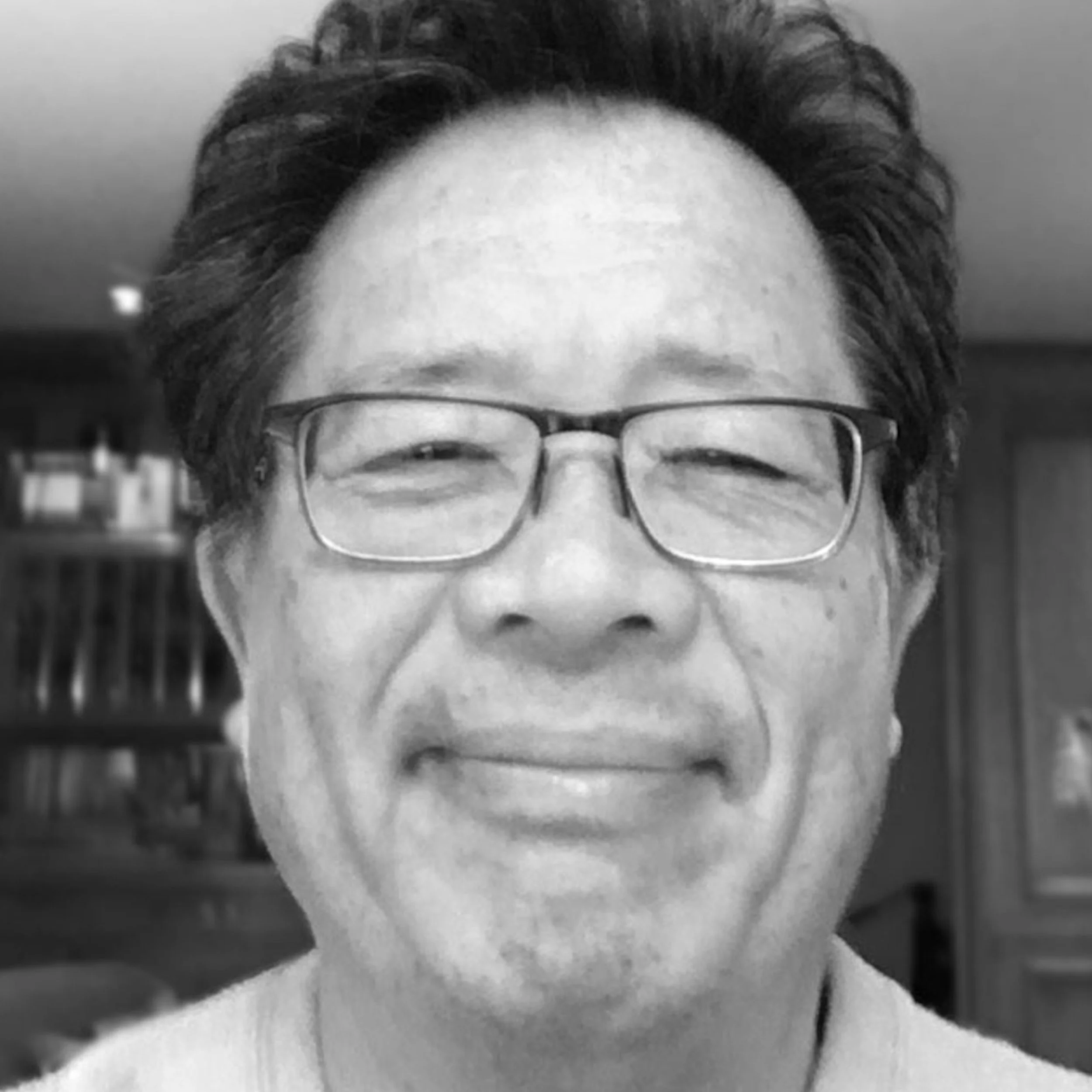 A close-up black and white photo of a smiling man with glasses and dark hair in an indoor setting.