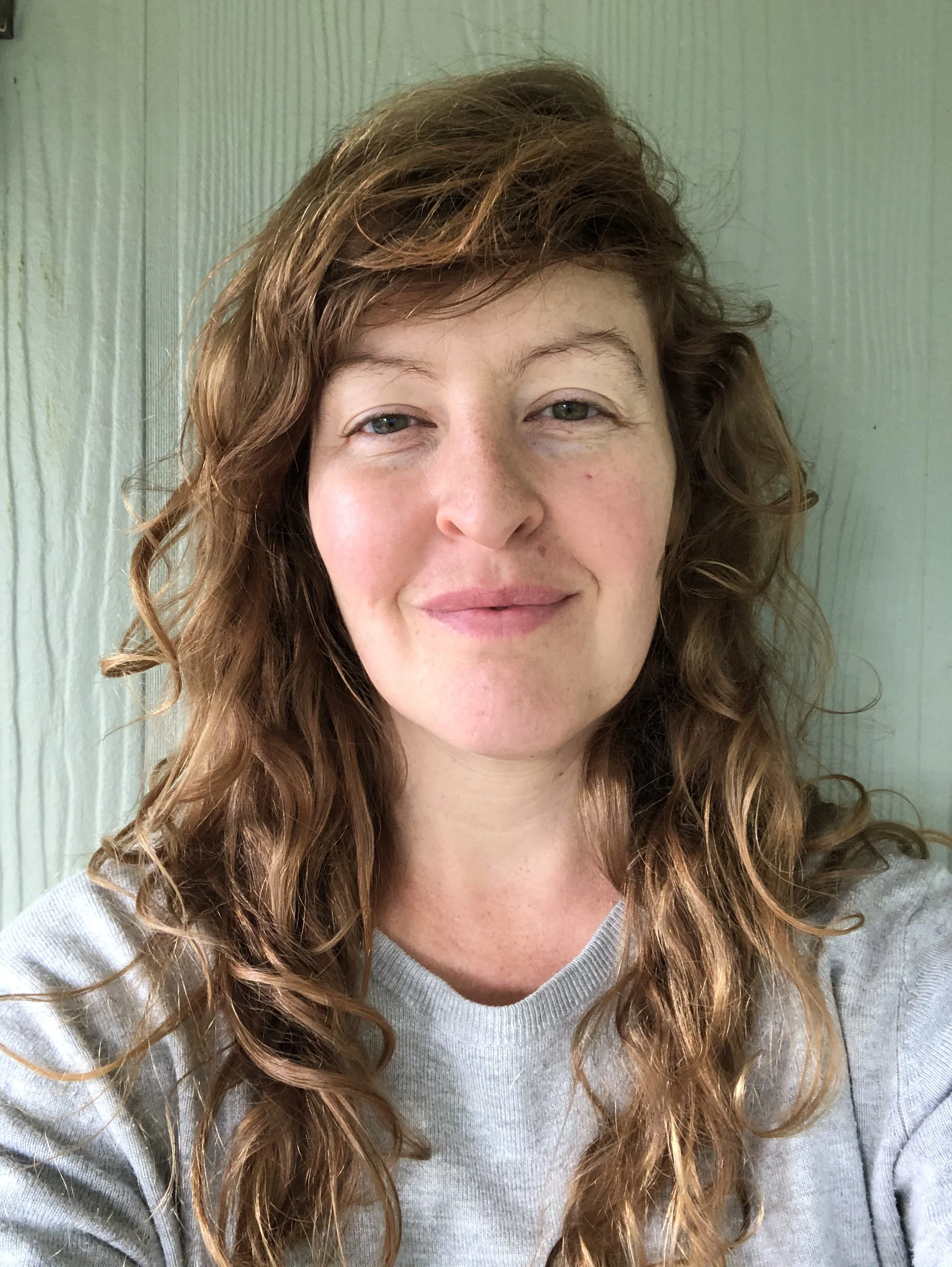 A woman with light skin and brown hair tied up in a bun, smiling, standing in front of three framed artworks of anatomical hearts with colorful artistic designs.