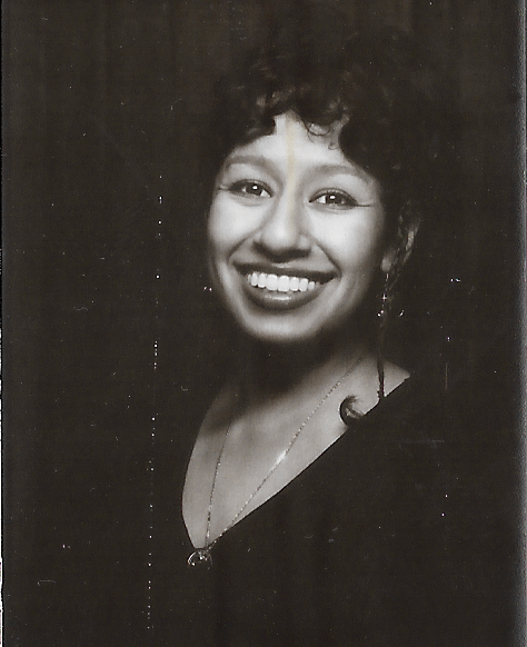 A young woman with short, curly black hair wearing a maroon sweatshirt, gold jewelry, and earrings, resting her chin on her hand and looking into the camera with a neutral expression against a plain white background.