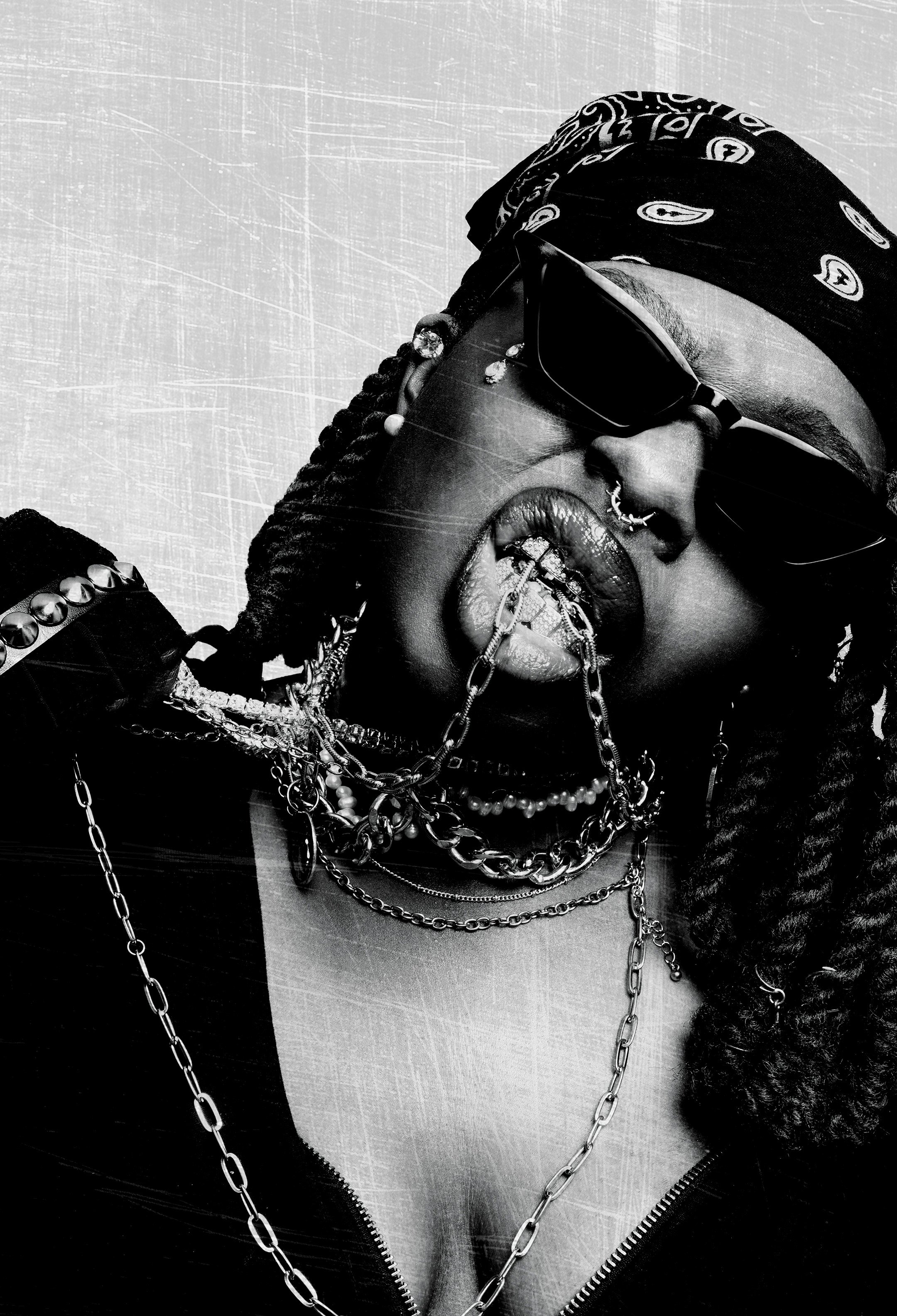 Black and white photograph of a person wearing dark sunglasses, with multiple necklaces and chain jewelry, a headscarf with patterns, and a jacket with metal studs.