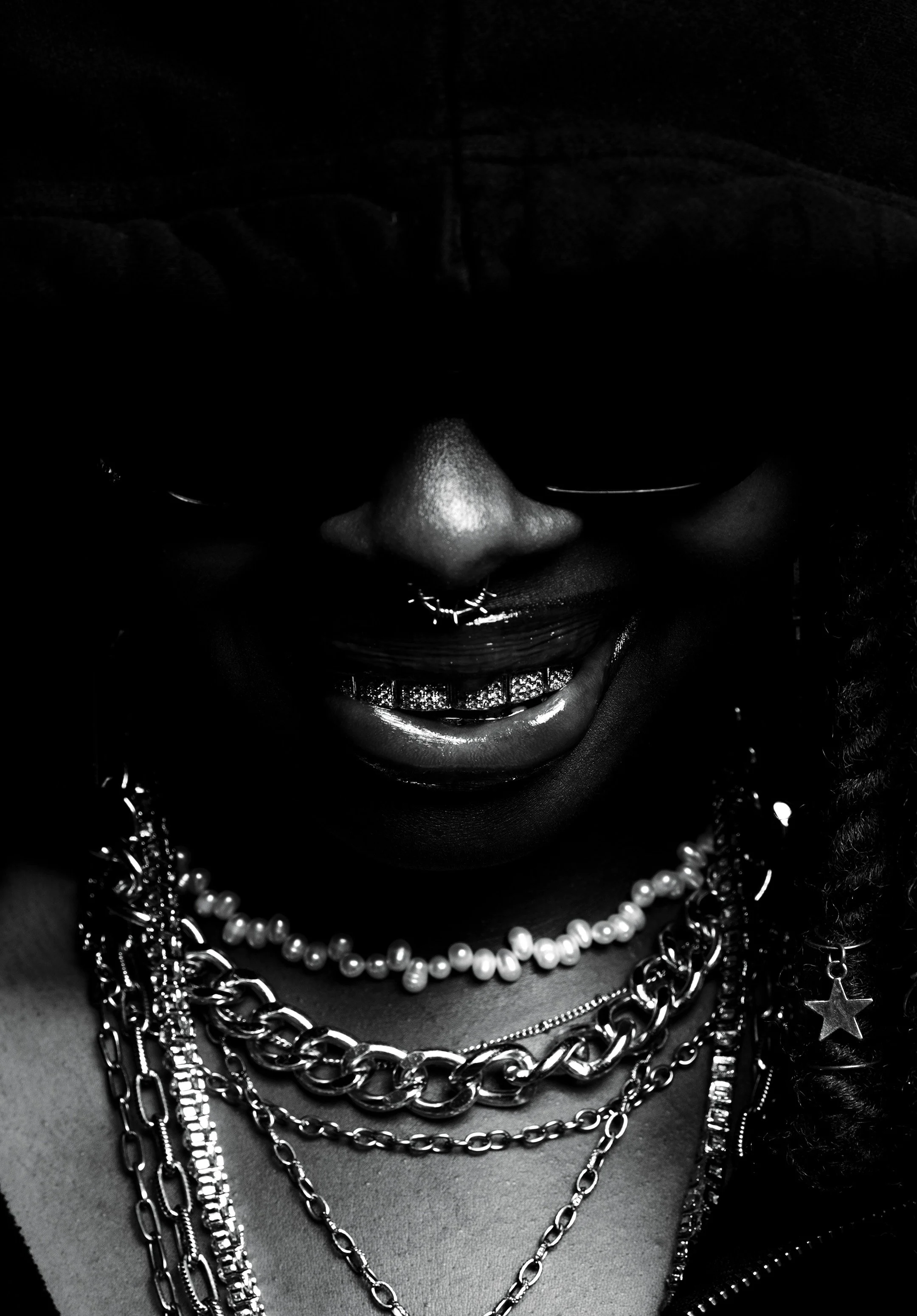 Close-up black and white photograph of a person's face with multiple necklaces, sunglasses, and a cap. The person is smiling slightly, showing a gold grill on the teeth and a septum piercing.