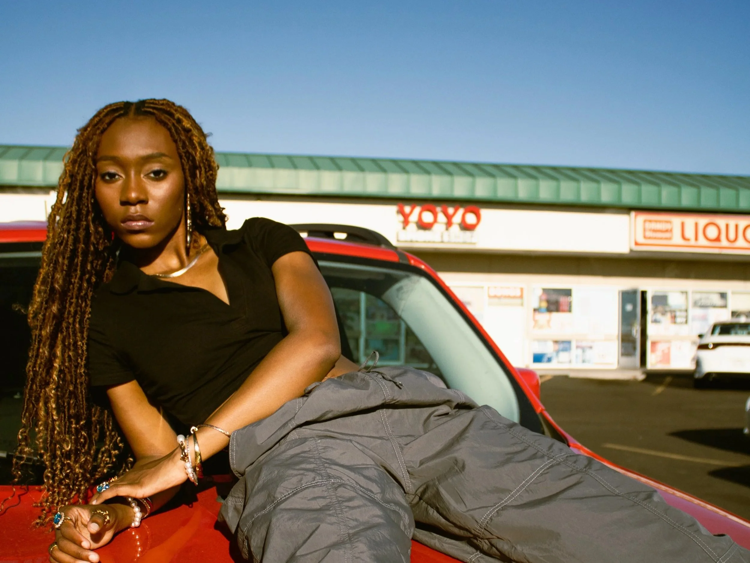 A woman with long braids sitting on the hood of a red car. She has a serious expression and is wearing bracelets and a black top. In the background, there is a store with a sign that reads 'YOYO' and other signs for liquor and convenience store.