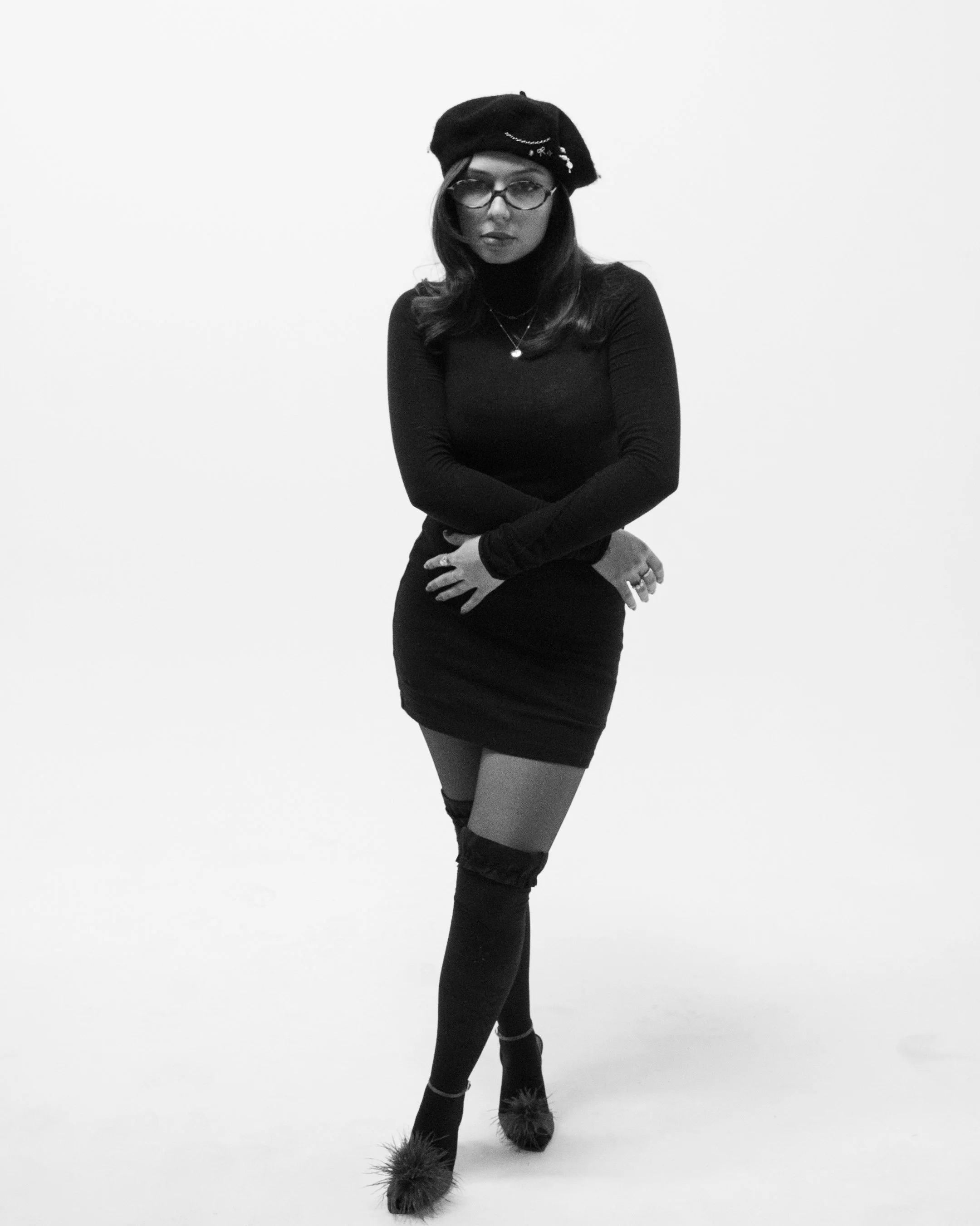A young woman dressed in black, wearing a beret with embellishments, glasses, a turtleneck, and thigh-high furry boots, standing against a plain white background.