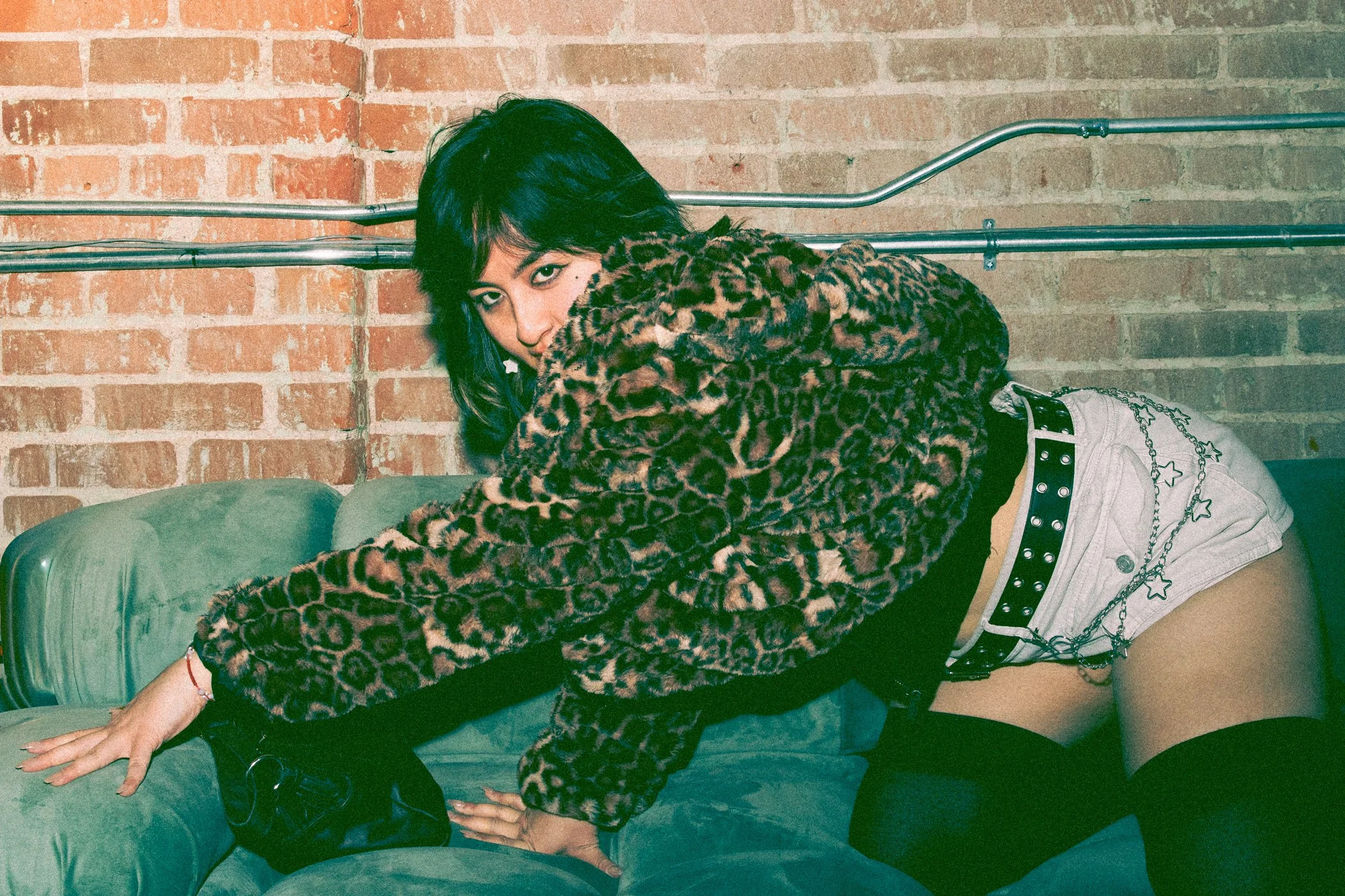 A woman with dark hair and leopard print jacket leaning over a green couch against a brick wall, posing with her head tilted and looking at the camera.