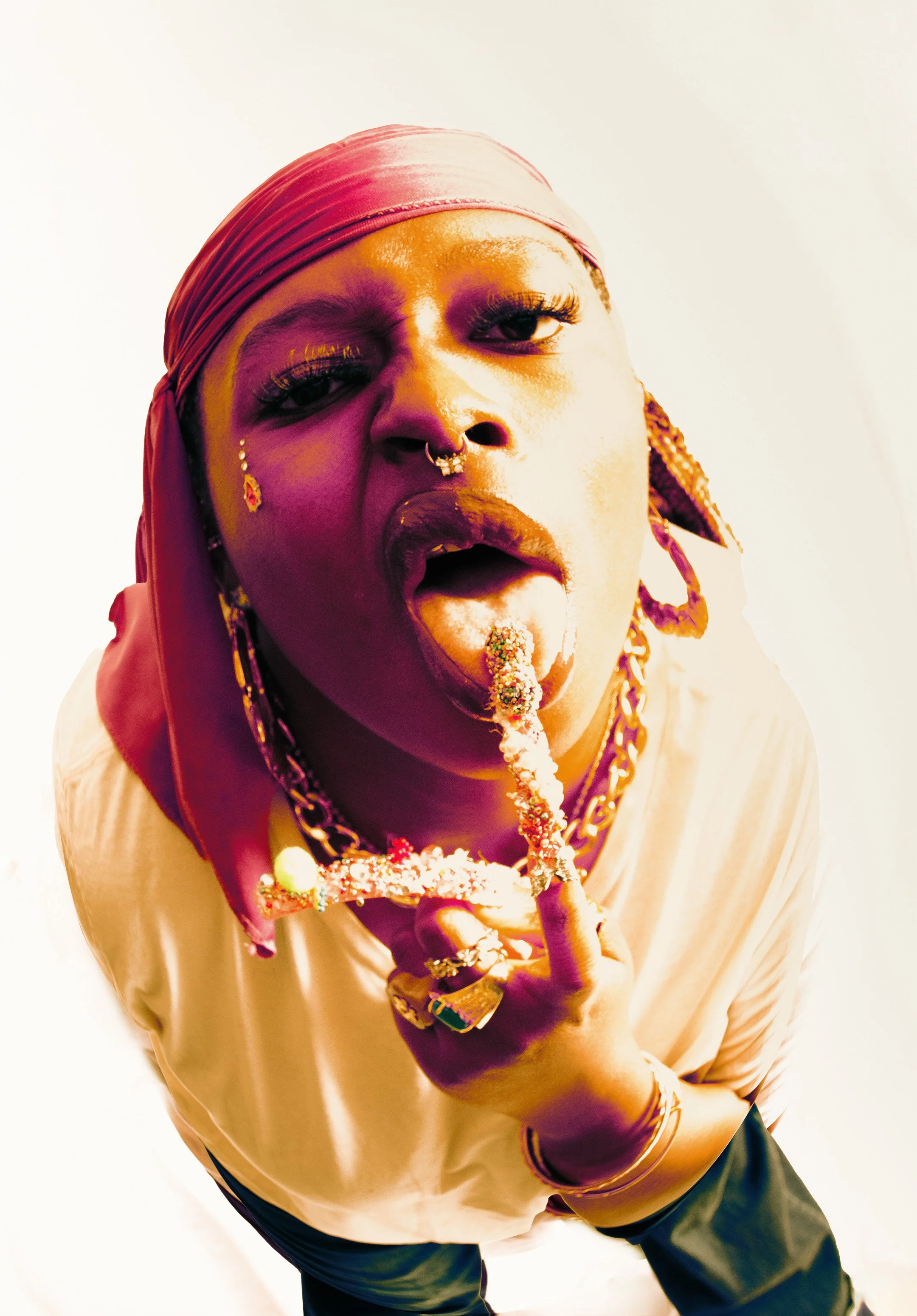 Person with a pink headscarf and gold jewelry, sticking out their tongue with their long acrylic nail
