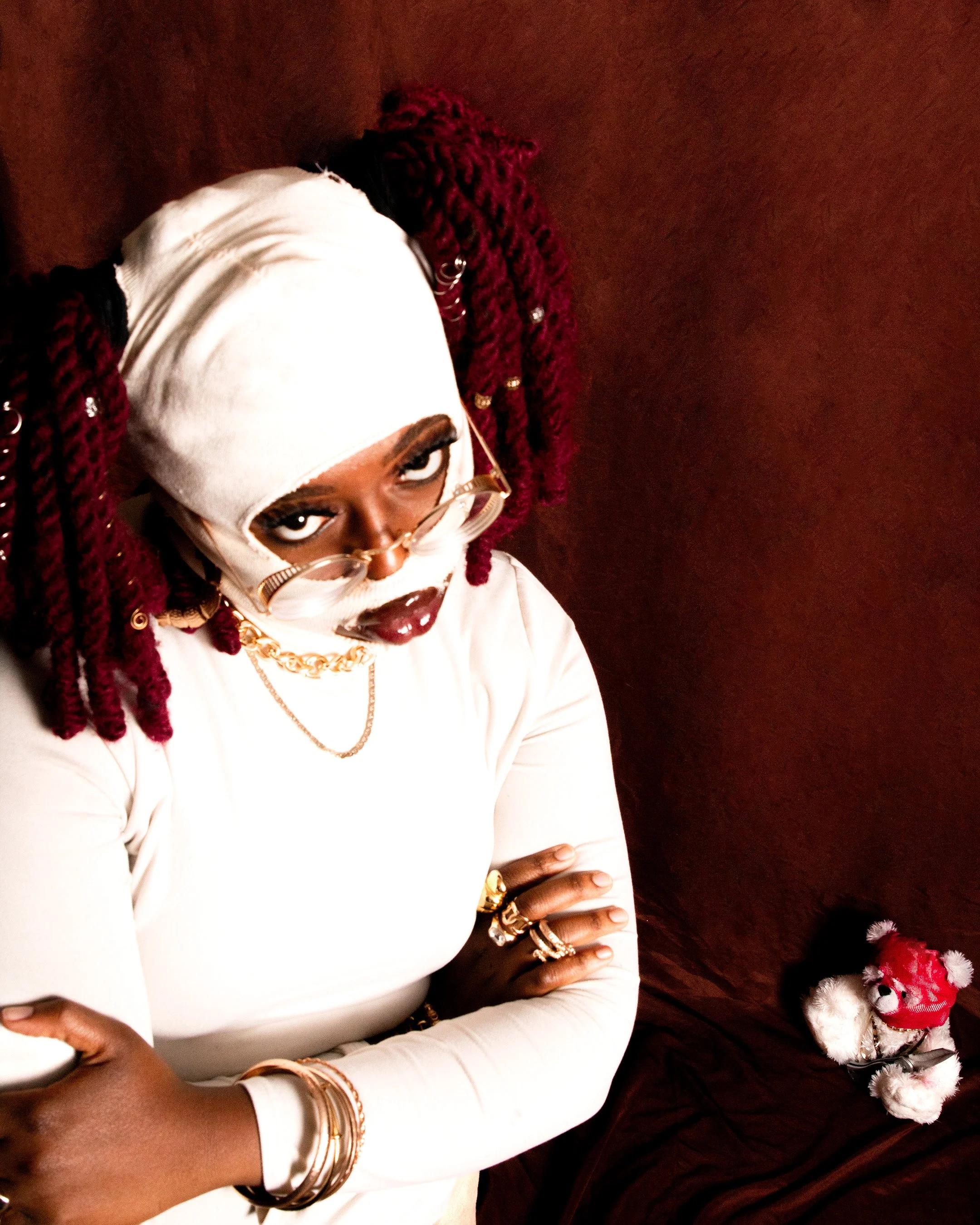 A woman with dark skin and red dreadlocks wearing large sunglasses, gold jewelry, a white long-sleeve shirt, and a beige headscarf, leaning against a dark background with a small stuffed teddy bear dressed as Santa Claus at the bottom right of the im