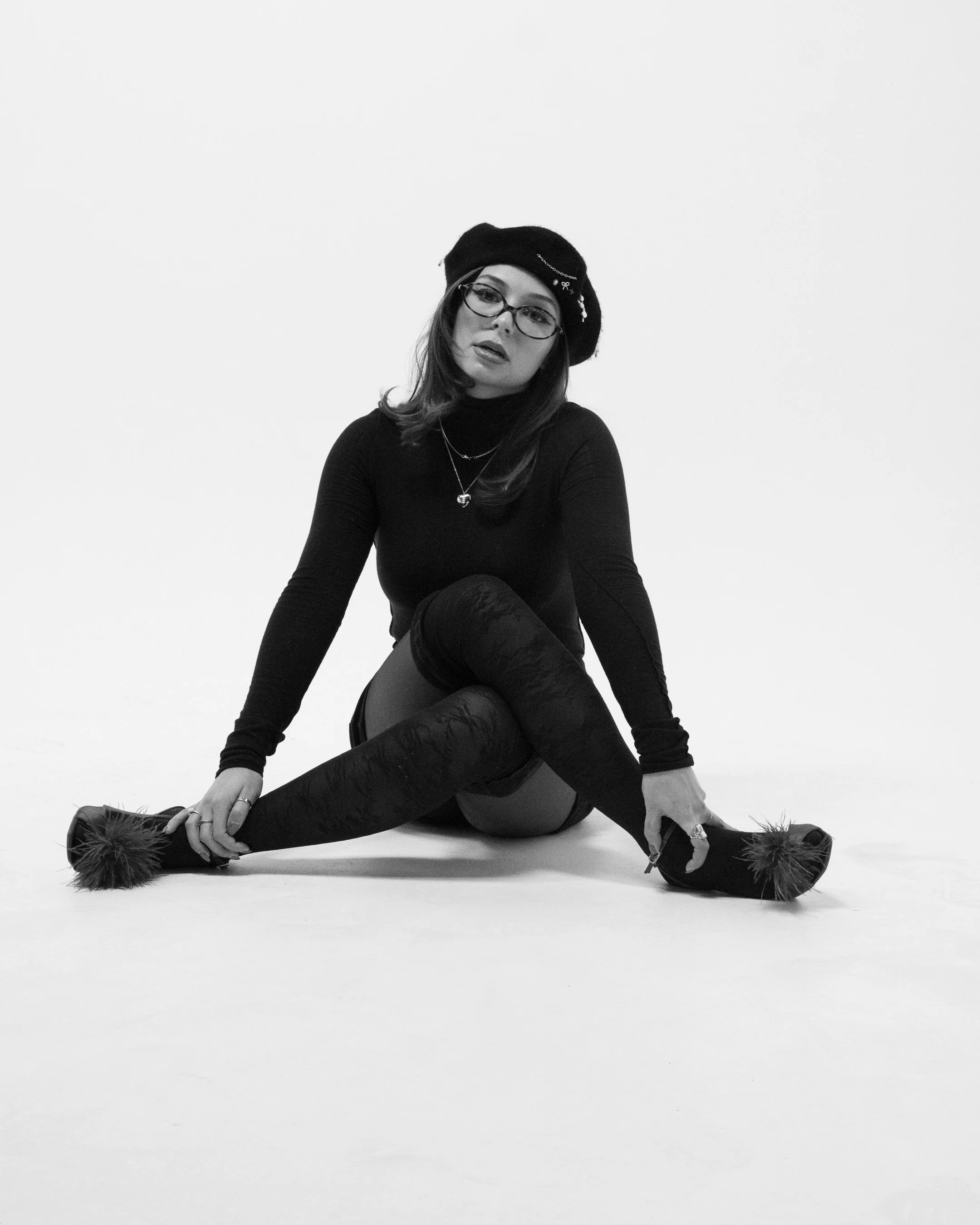 A monochrome photo of a woman with glasses and a beret, sitting on the floor with her legs extended and crossed, surrounded by a plain white background.