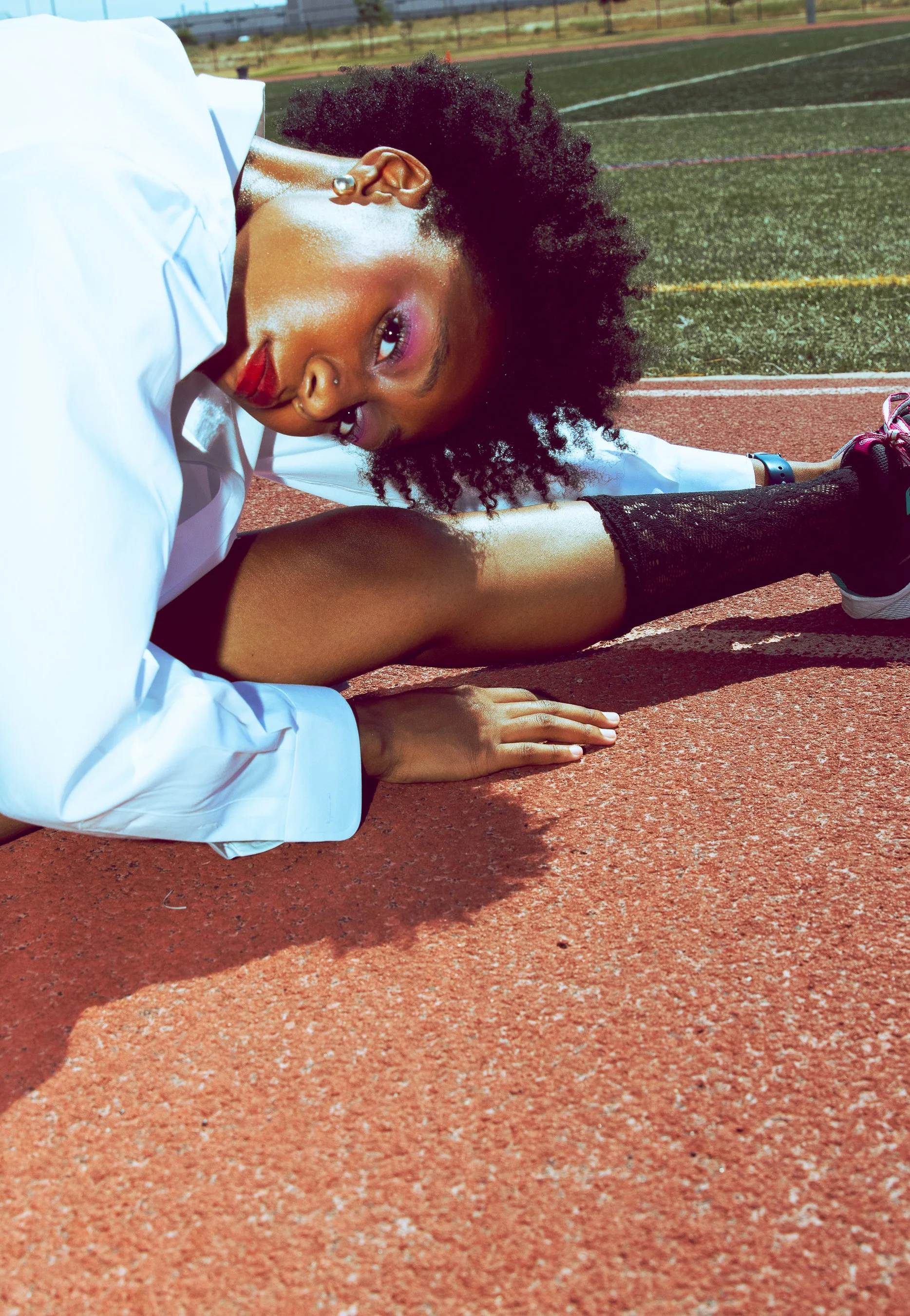 A young woman with curly hair lying on a running track, stretching with her arms extended and her head resting on the ground, wearing athletic gear and a gym tracker on her wrist.