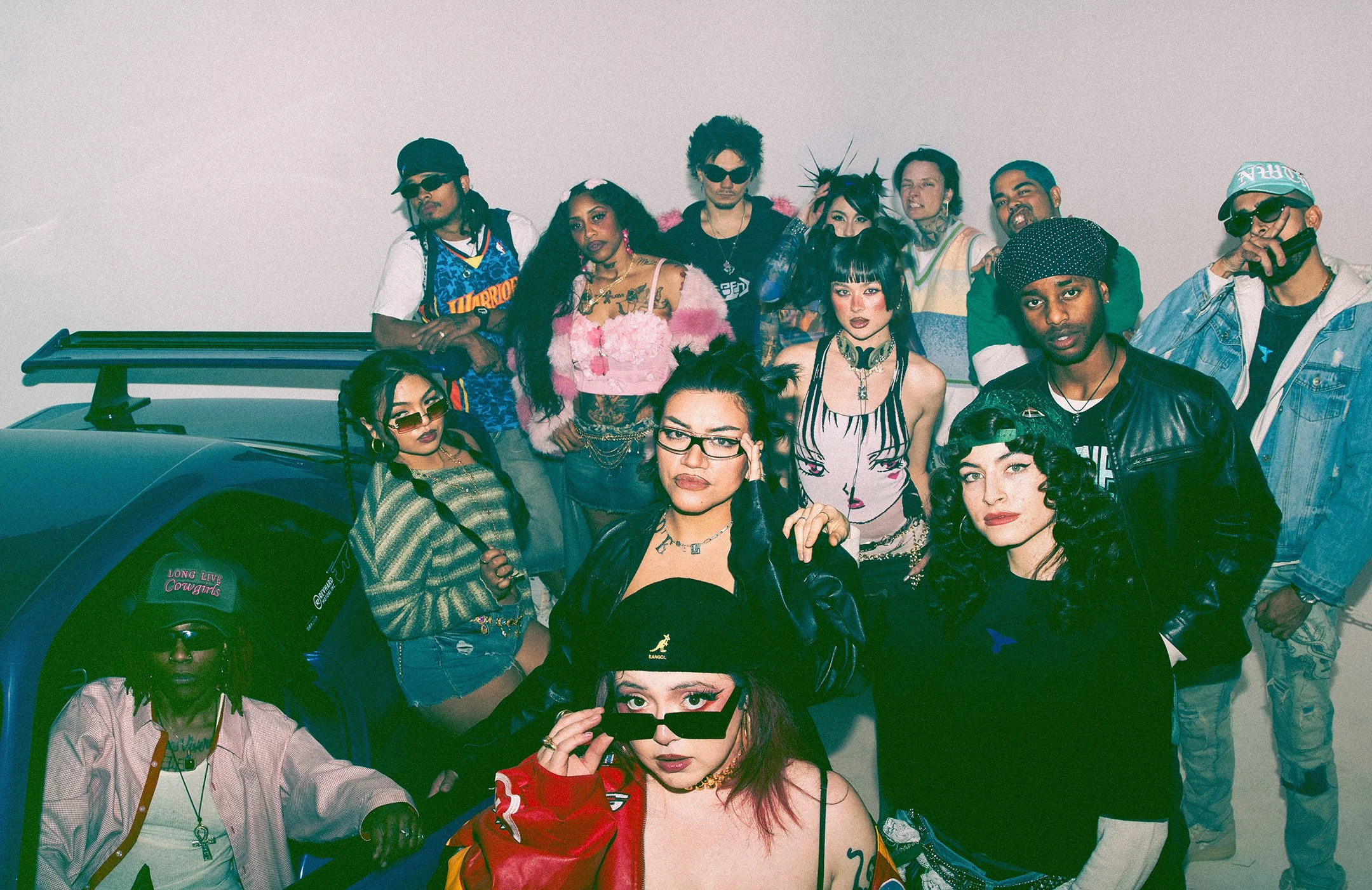Group of diverse young people posing in front of a plain white wall, some sitting in a car, dressed in colorful and trendy clothing, with various accessories, sunglasses, and expressive hairstyles, creating a vibrant, edgy atmosphere.