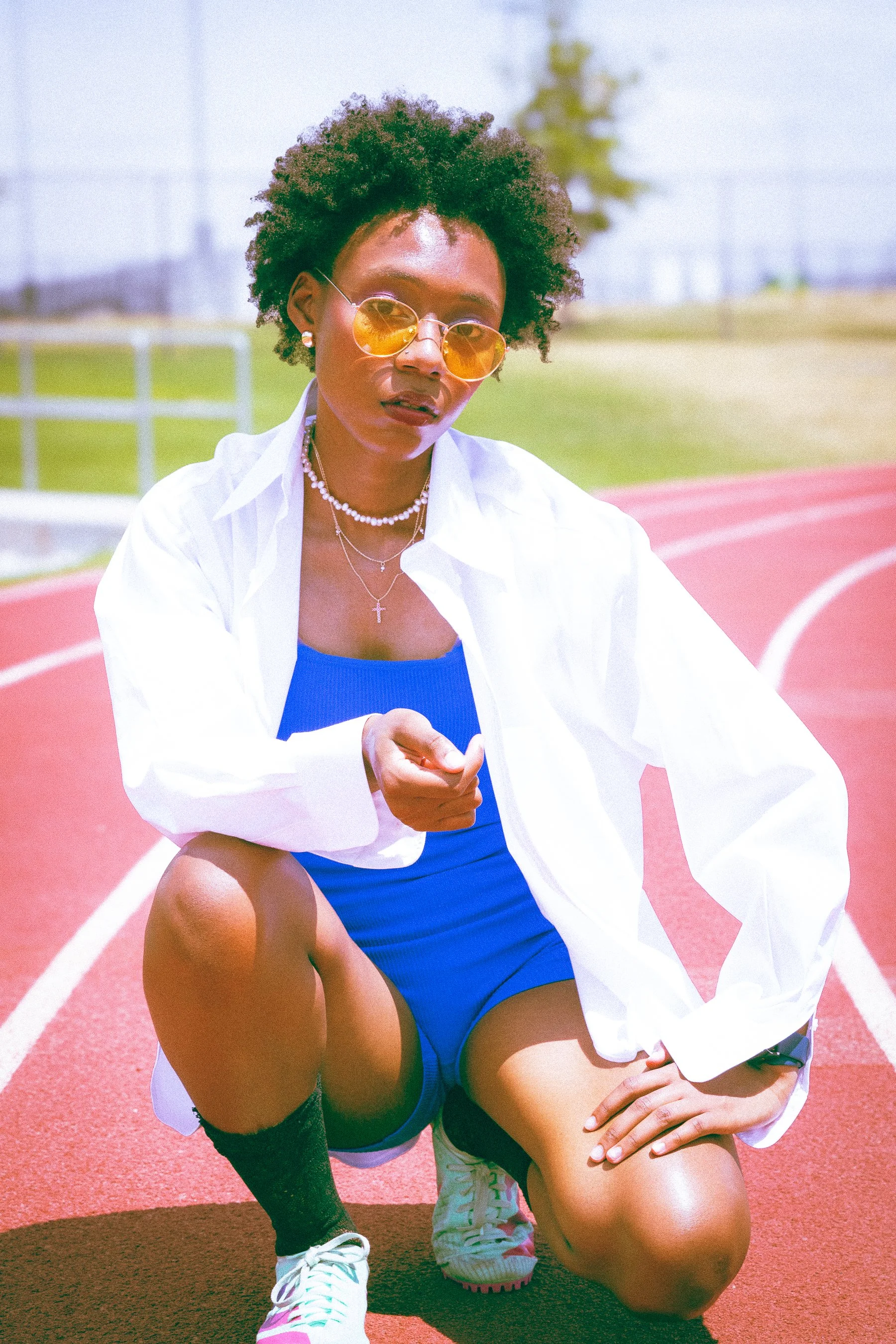 A woman with an afro hairstyle wearing sunglasses, layered necklaces, a white shirt, and blue athletic wear, crouching on a red track field with one knee on the ground, in an outdoor sports facility with a green field and trees in the background.