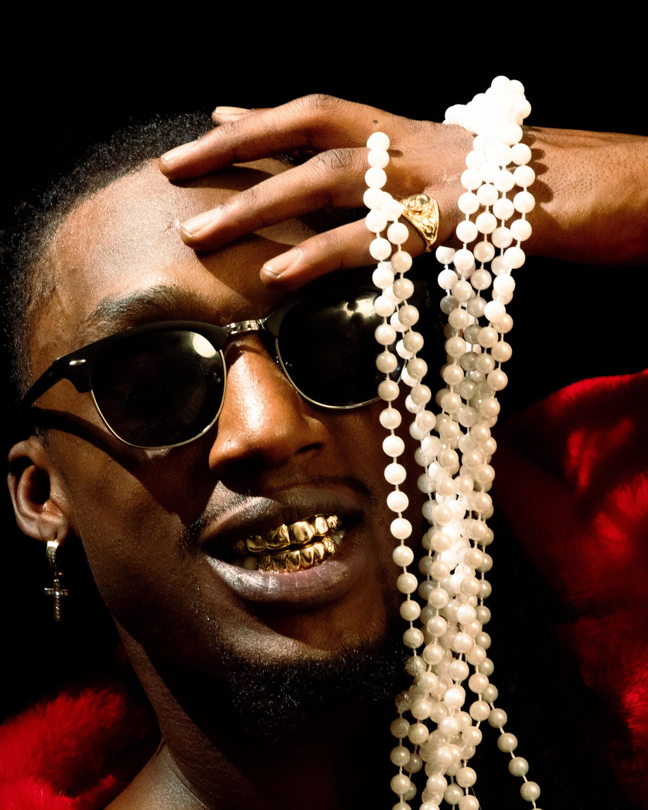 Close-up of a person with dark skin wearing black sunglasses, gold teeth, a gold ring, a cross earring, and a red fur coat, holding white pearl necklaces and with short black hair.