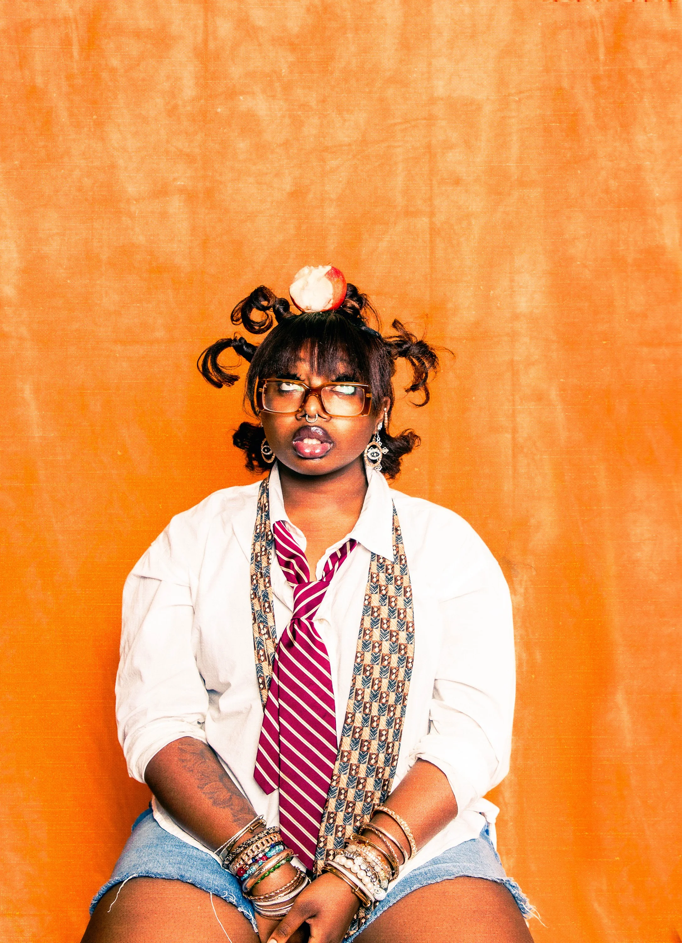 A young woman with dark skin and curly hair styled into pigtails topped with a half-eaten red apple. She wears large glasses, a septum piercing, bold lipstick, earrings, and multiple bracelets. She's dressed in a white shirt, a striped tie, and denim