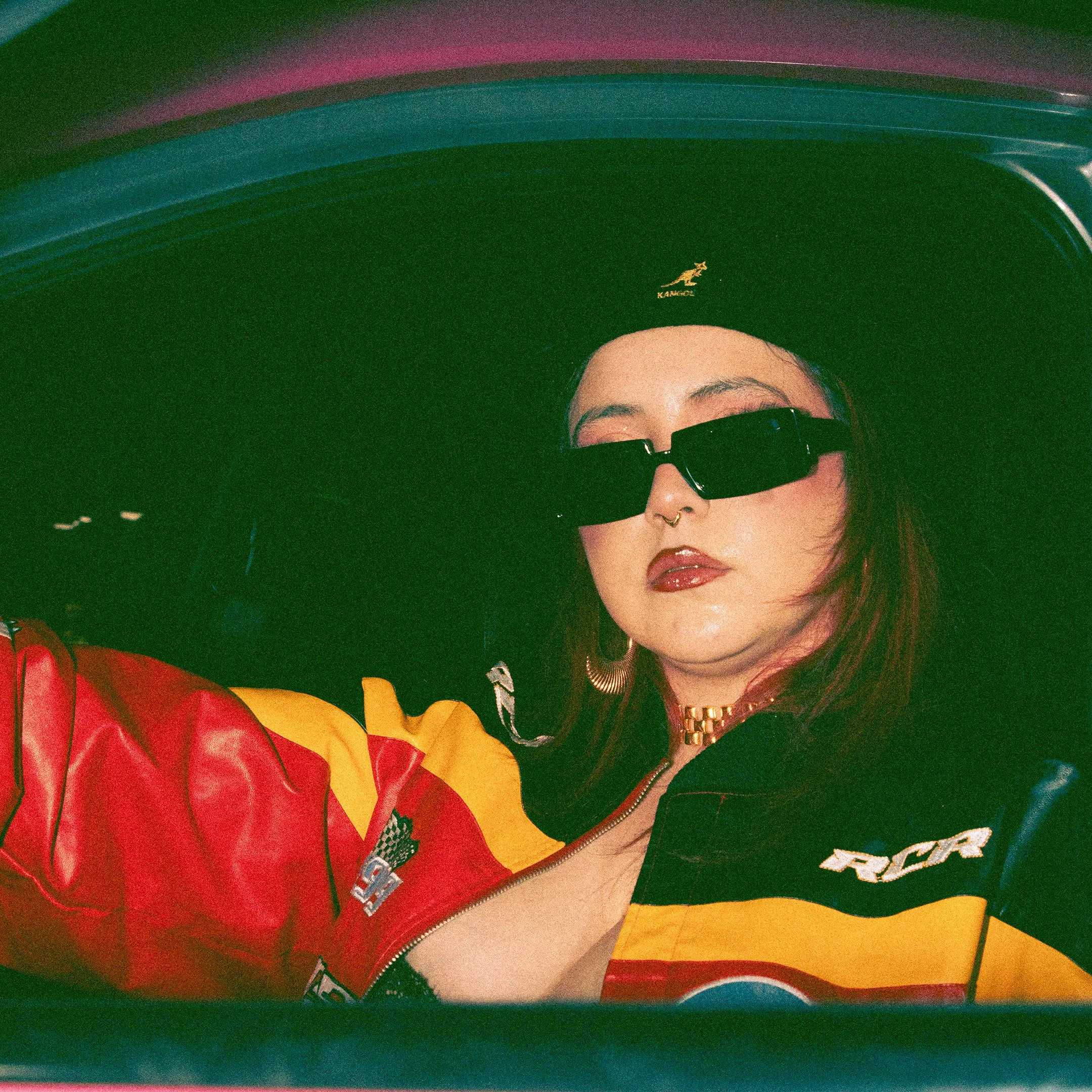 Part of a person's face, including eyes, nose, lips, wearing sunglasses and a black jacket, with a colorful jacket visible, behind a large, shiny, red and yellow balloon.