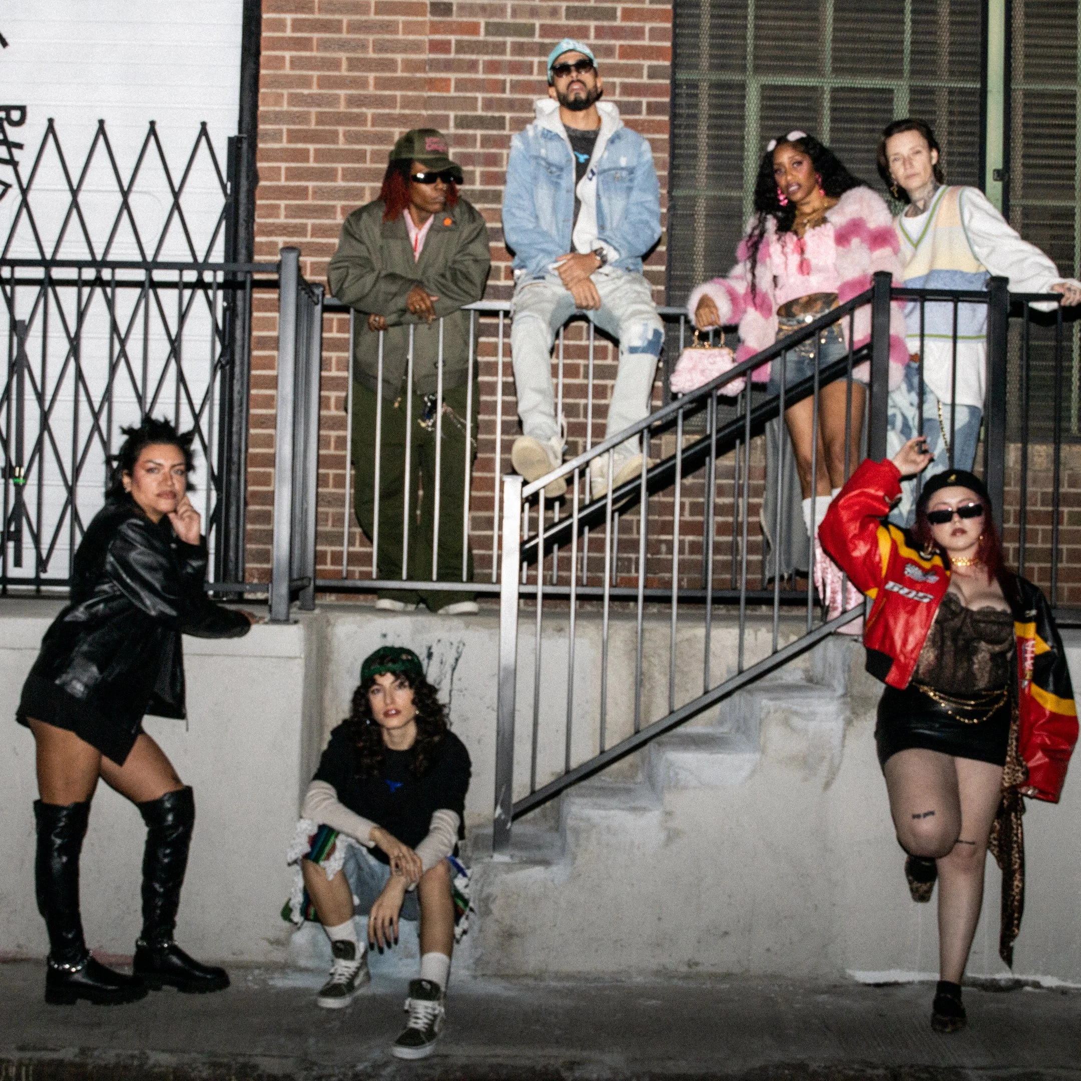 Group of five diverse young adults posing on a concrete staircase outside a building with brick wall and window. Some are sitting on the stairs and some are standing, dressed in casual fashionable clothing.