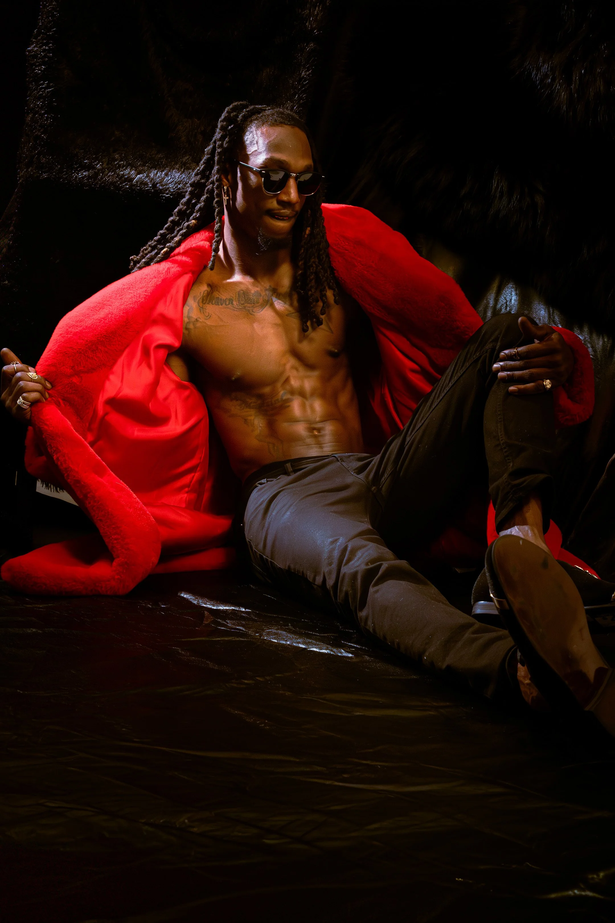 A shirtless man with long dreadlocks and sunglasses sitting against a black background, wearing a red coat draped over his shoulders.