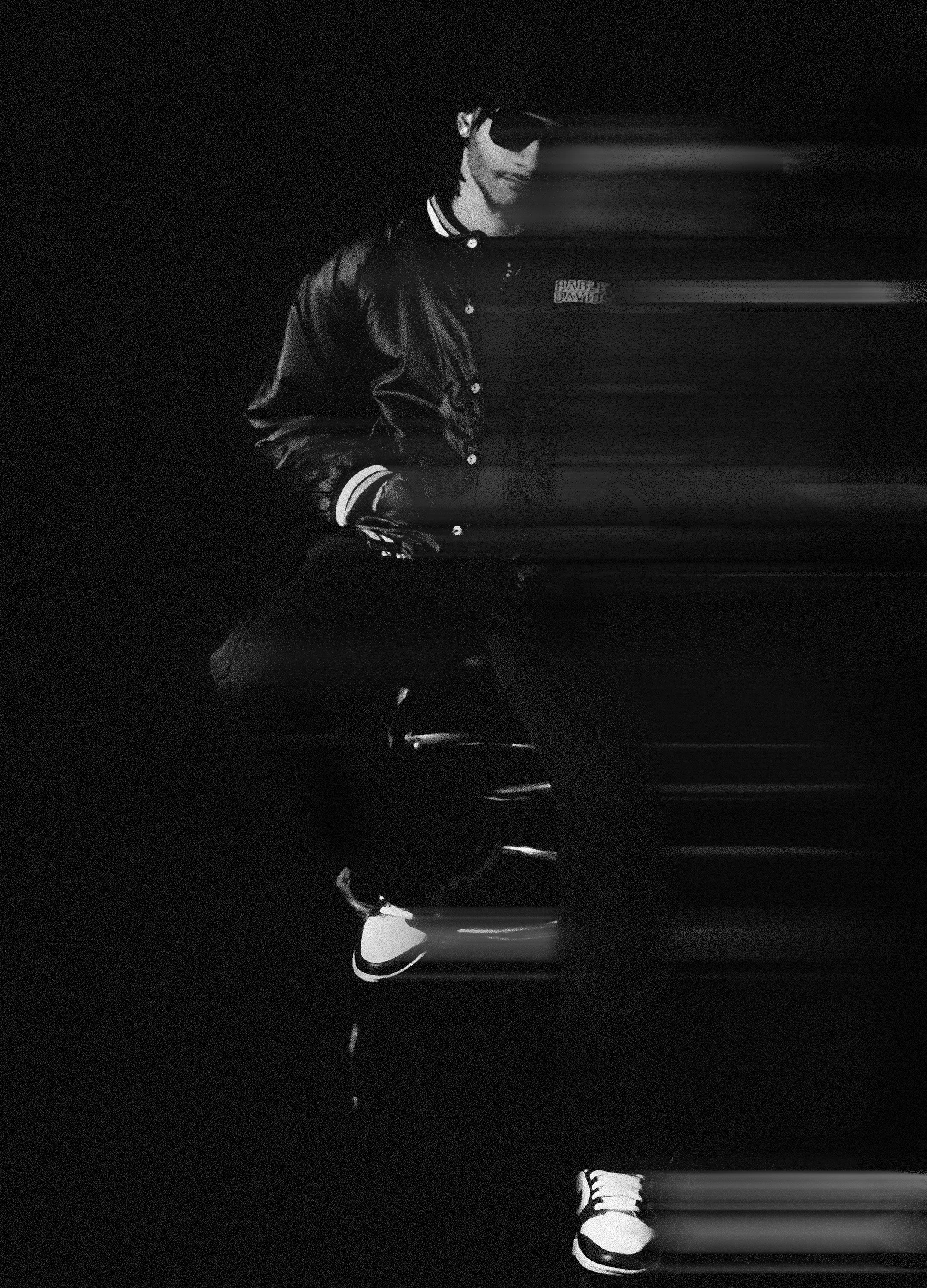 A young man wearing a leather jacket, striped shirt, sneakers, sunglasses, and headphones, standing outdoors at night with motion blur effects.