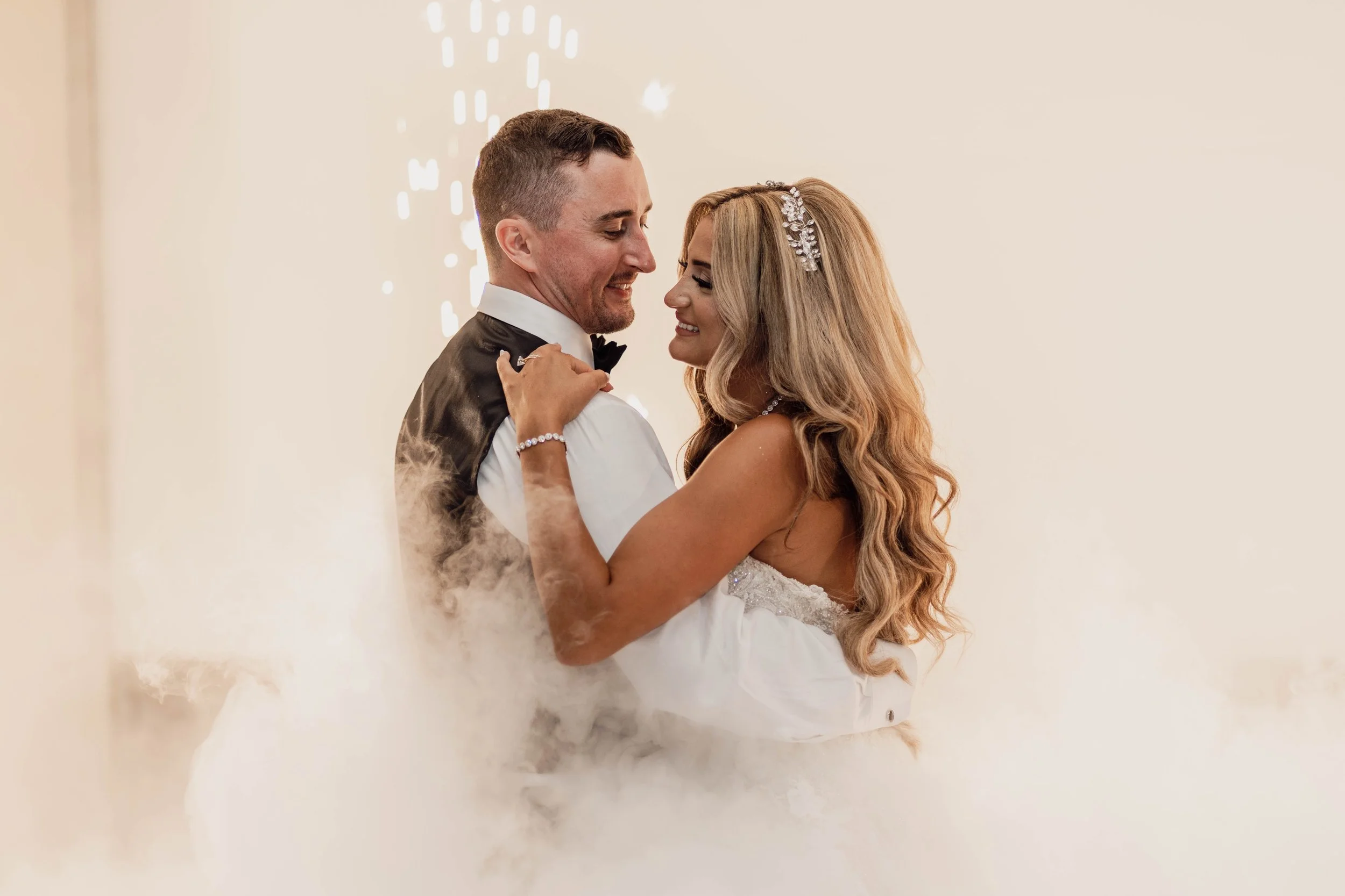 Bride and groom dancing closely at their wedding, with bride wearing a strapless wedding dress and headpiece, and groom in a tuxedo with vest and bow tie, surrounded by a foggy atmosphere.