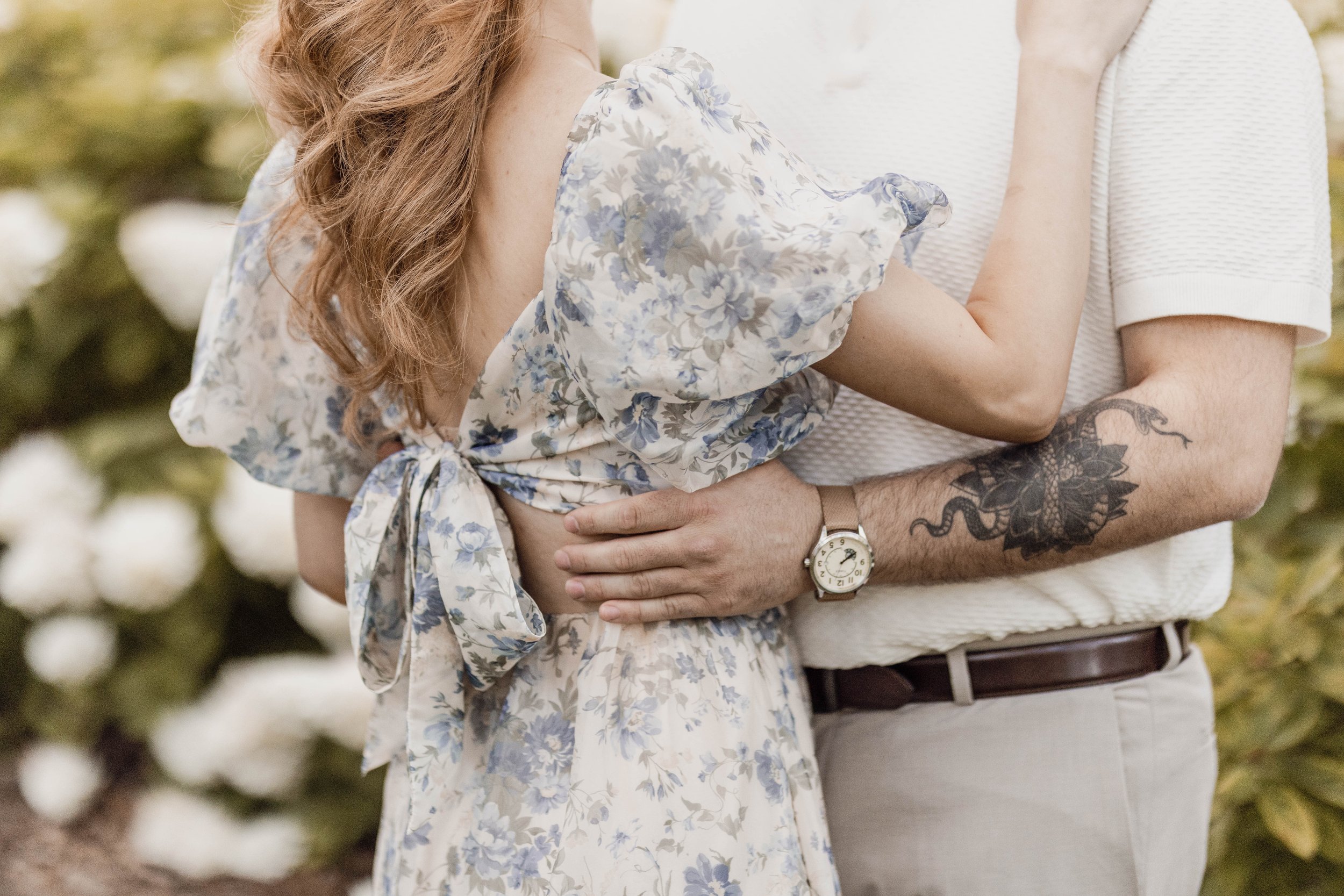 Close-up of a couple embracing outdoors, showing focus on their torsos and arms, with the woman's hand resting on the man's chest and the man’s hand around her waist, surrounded by greenery.