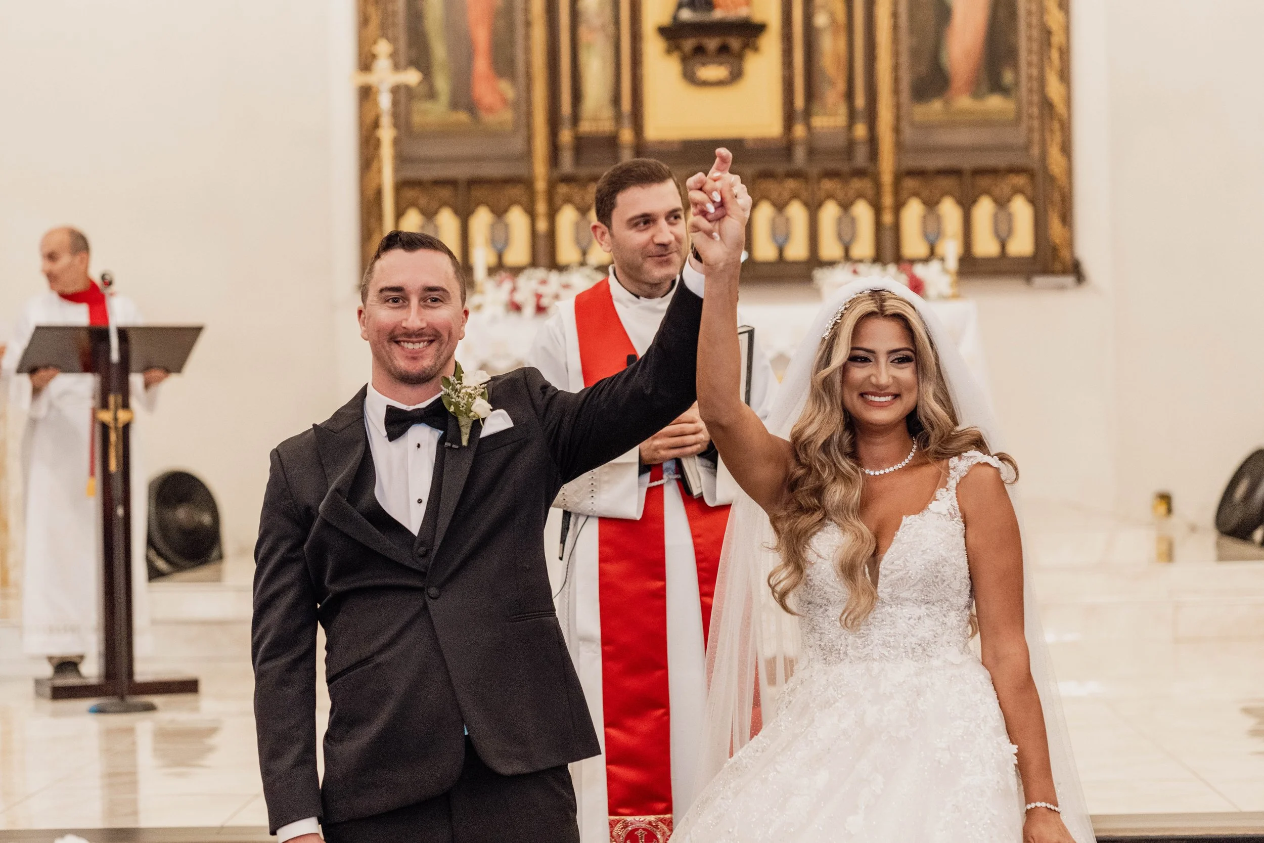 Newlywed couple holding hands raised in celebration during wedding ceremony inside a church with an officiant and a choir in the background.