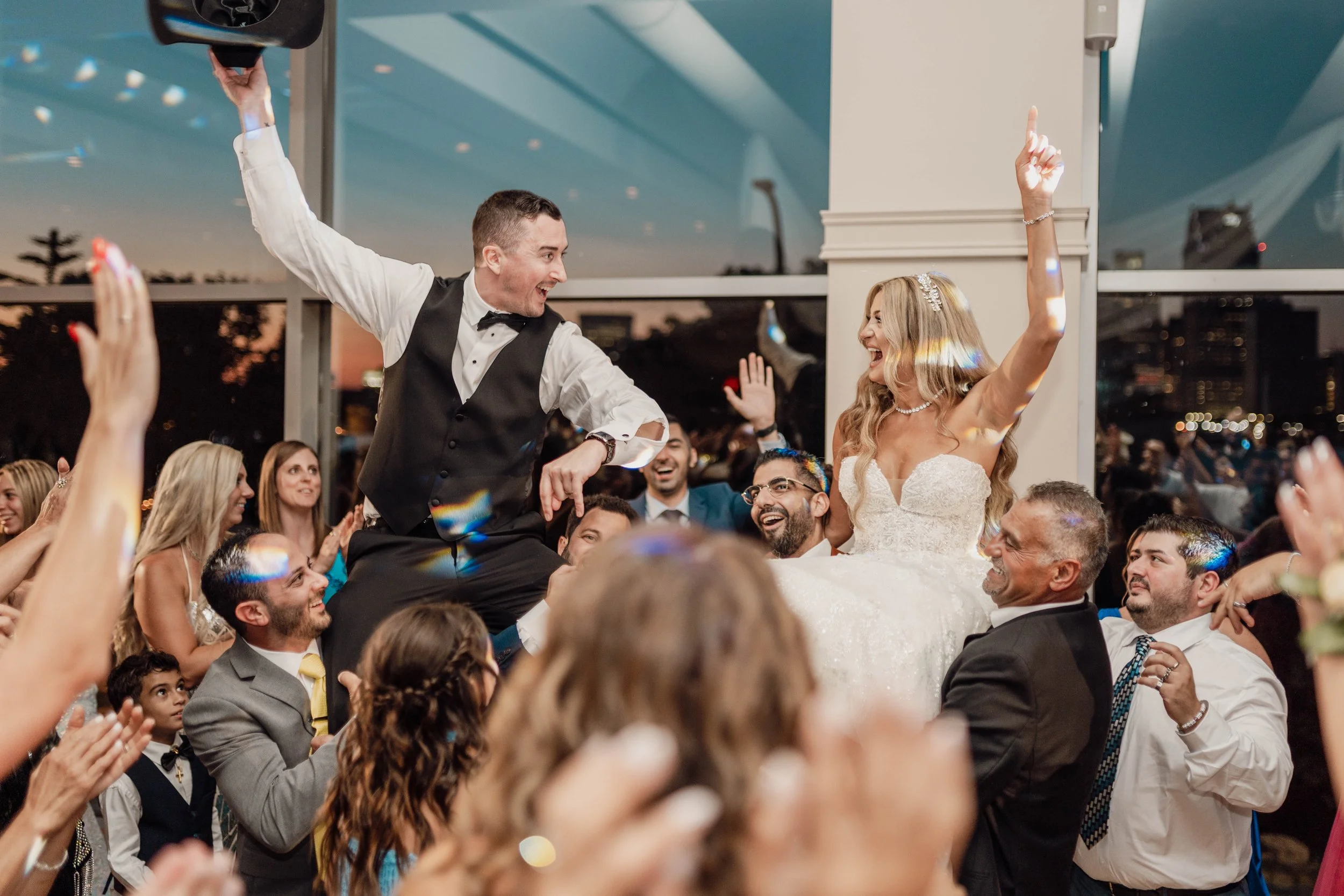Bride and groom on a wedding dance floor, the groom is sitting on the bride's lap, both smiling, while a crowd of wedding guests cheer around them, with a cityscape visible through large windows in the background.