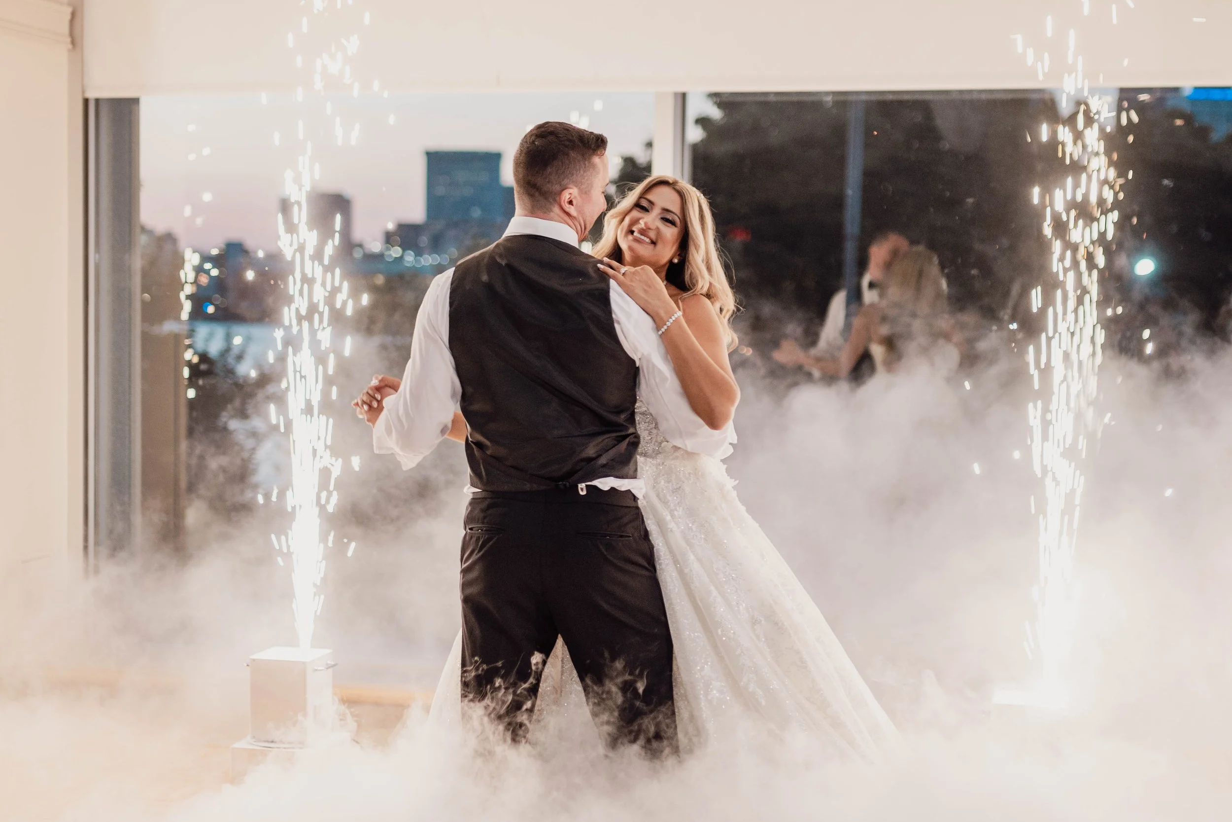 A bride and groom dancing together at their wedding reception, with sparklers and smoke effects in the background.