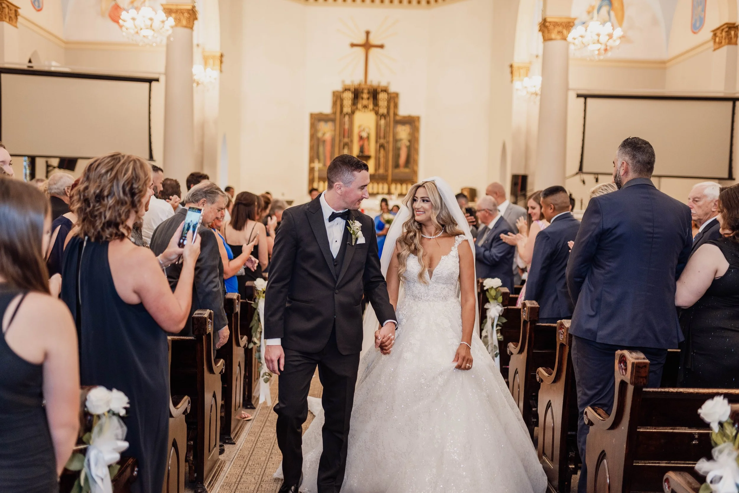 A bride and groom holding hands and smiling at each other during their wedding ceremony in a church, surrounded by seated guests, some taking photos.