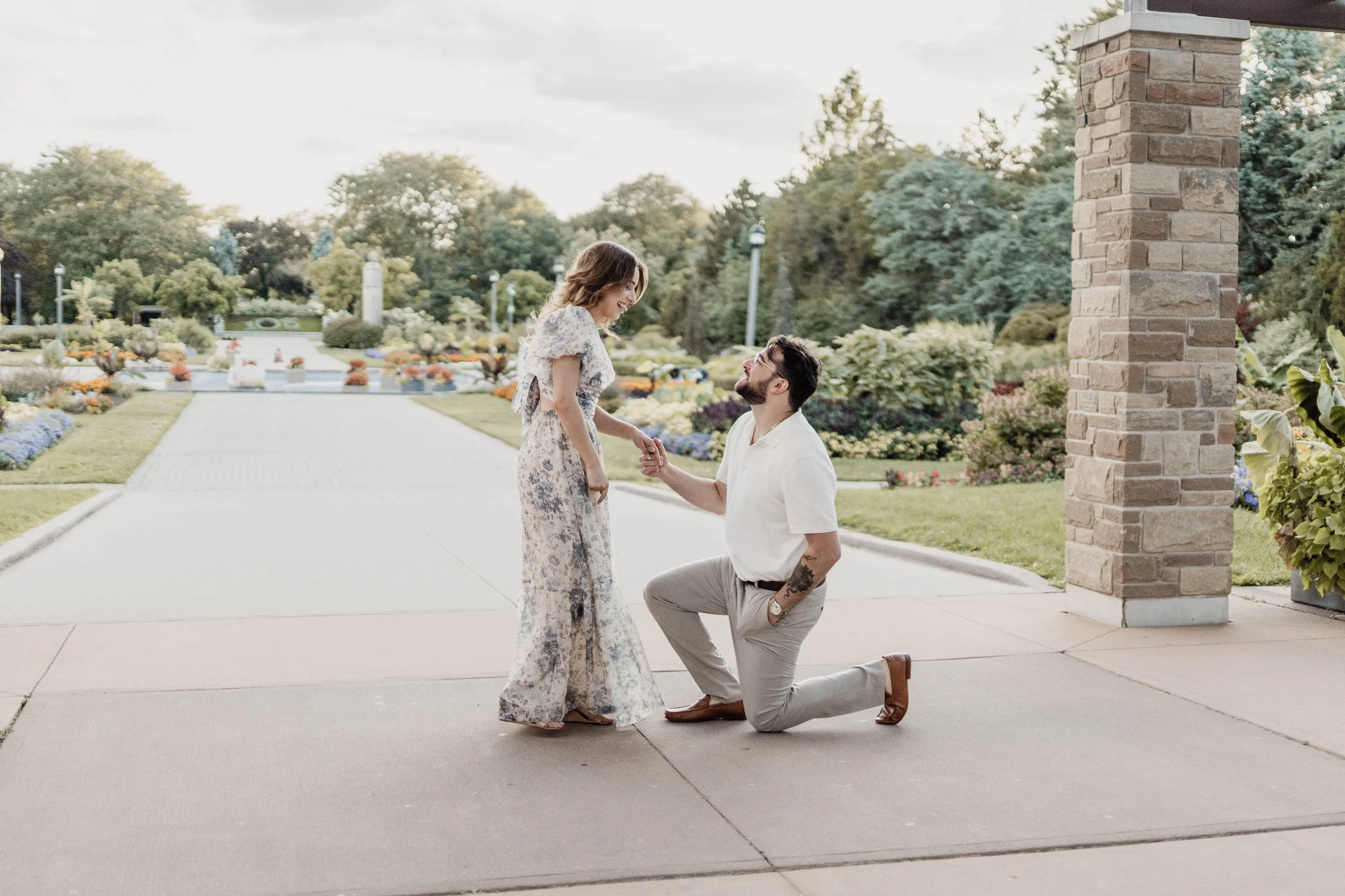 A man proposes marriage to a woman in a park with flowers, with the man kneeling on one knee and holding the woman's hand as she looks at him smiling.