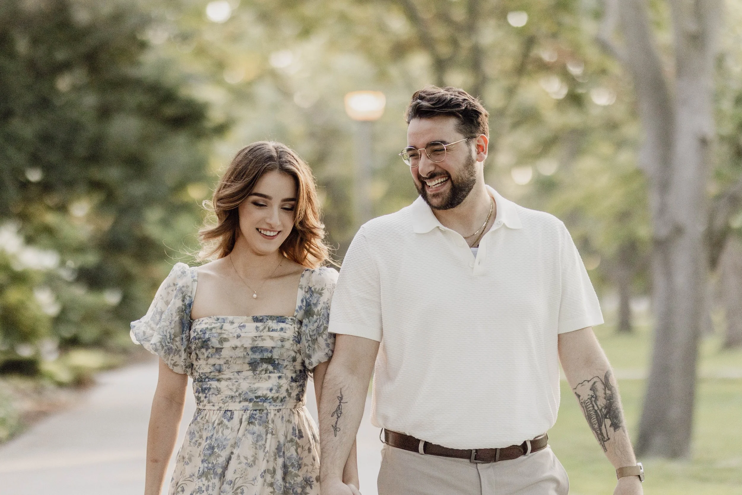 A young couple walking outdoors holding hands, smiling, with trees and a pathway in the background.