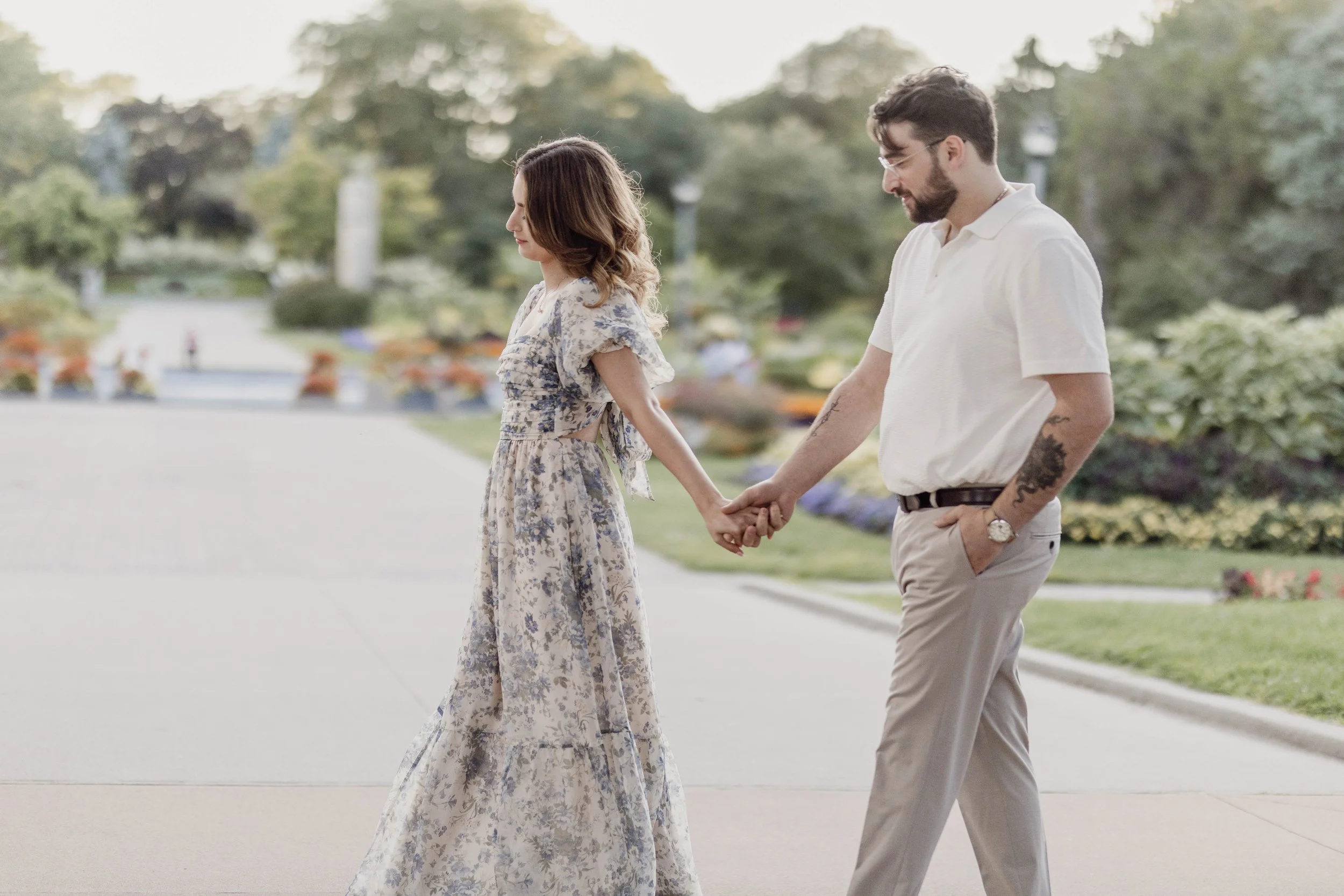 A young woman and man holding hands and walking outdoors in a park with colorful flowers and greenery.