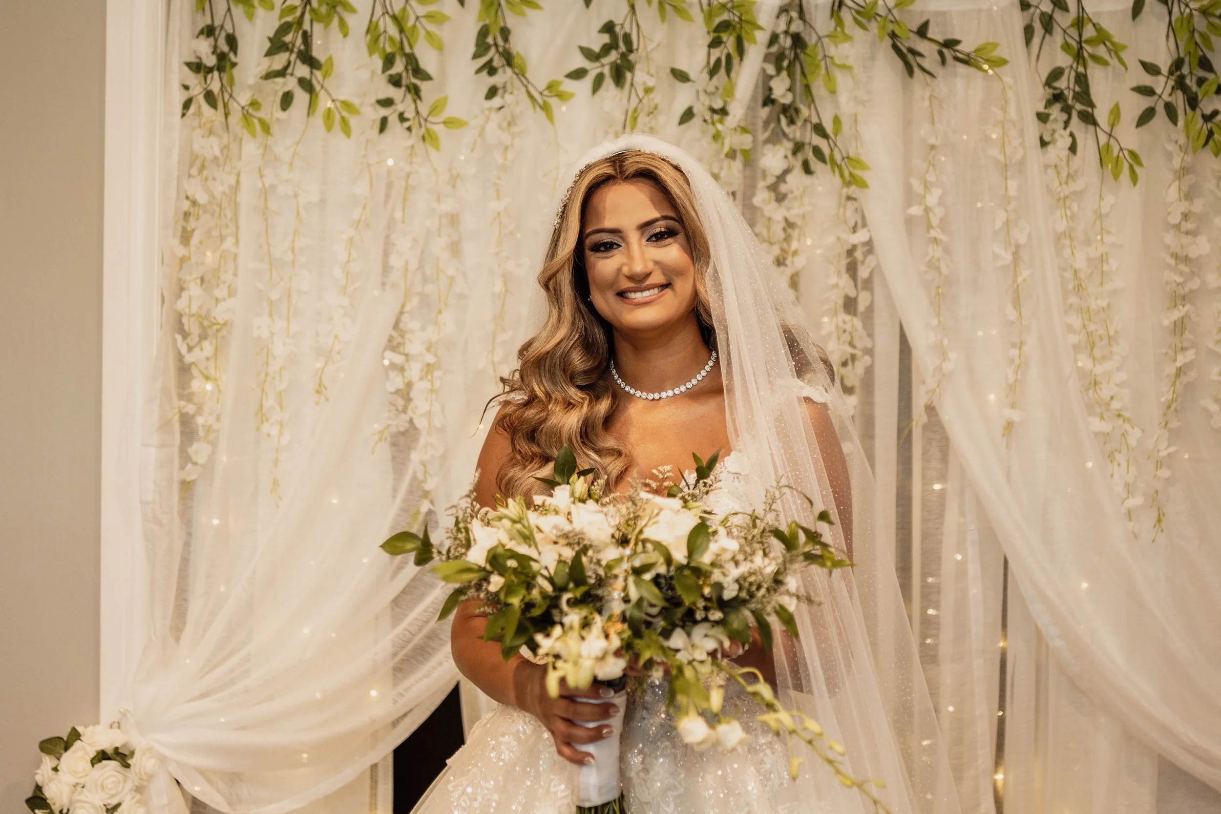 A bride with long, wavy hair, wearing a wedding dress and veil, standing in front of a floral backdrop holding a bouquet of white flowers and greenery, smiling at the camera.