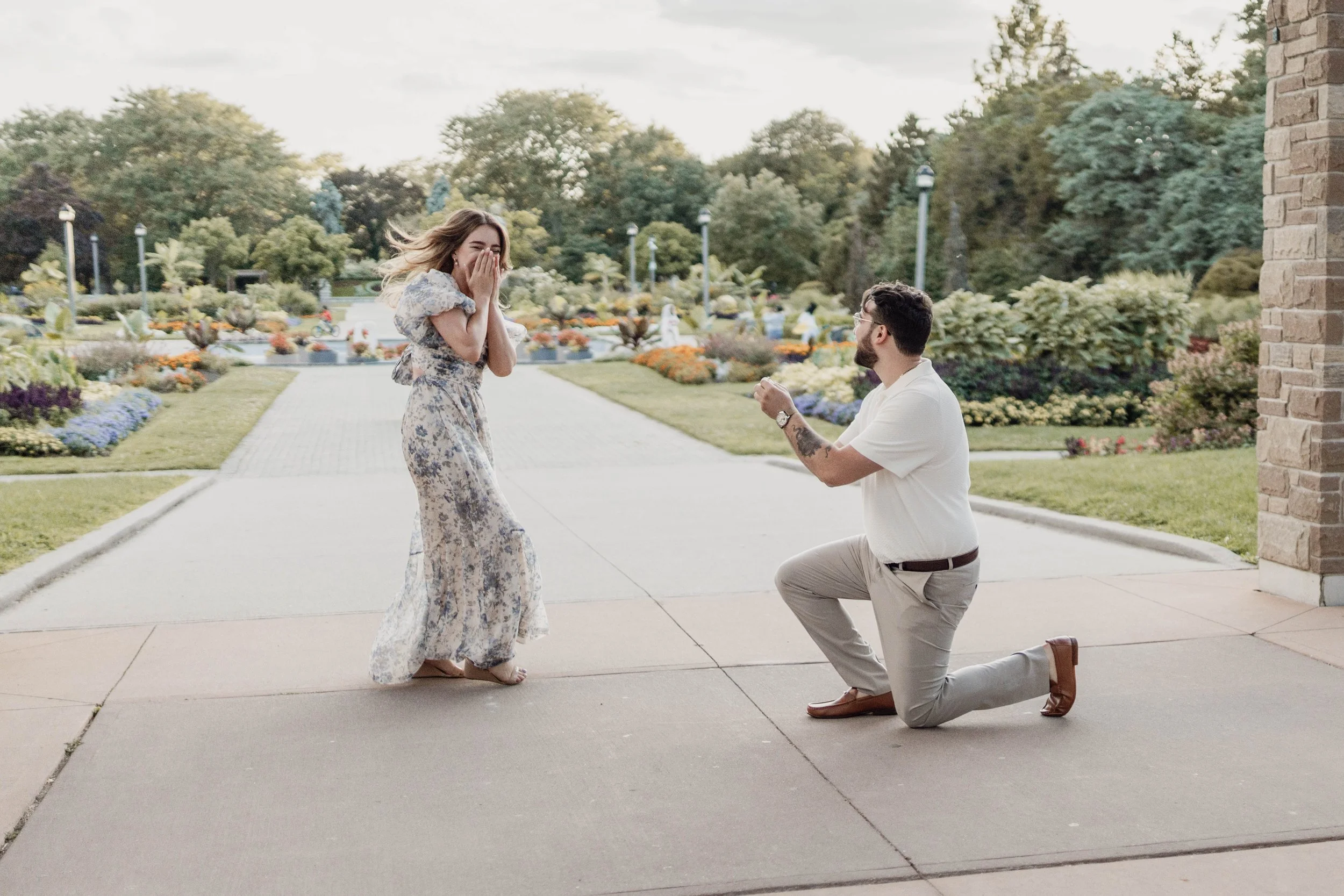 A man kneeling on one knee proposing to a woman in a garden with flower beds and trees, during daytime.