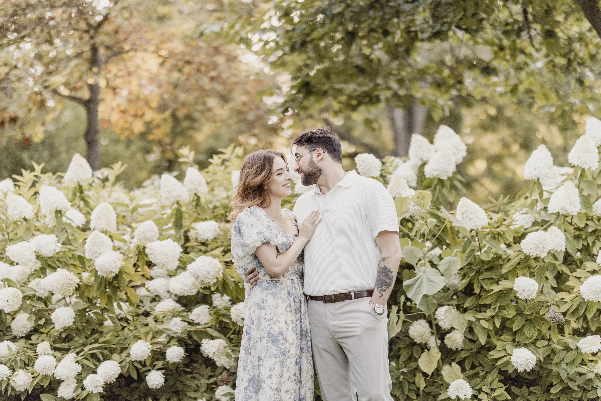 A young couple standing close together in front of blooming white hydrangea flowers outdoors, smiling and looking into each other's eyes.