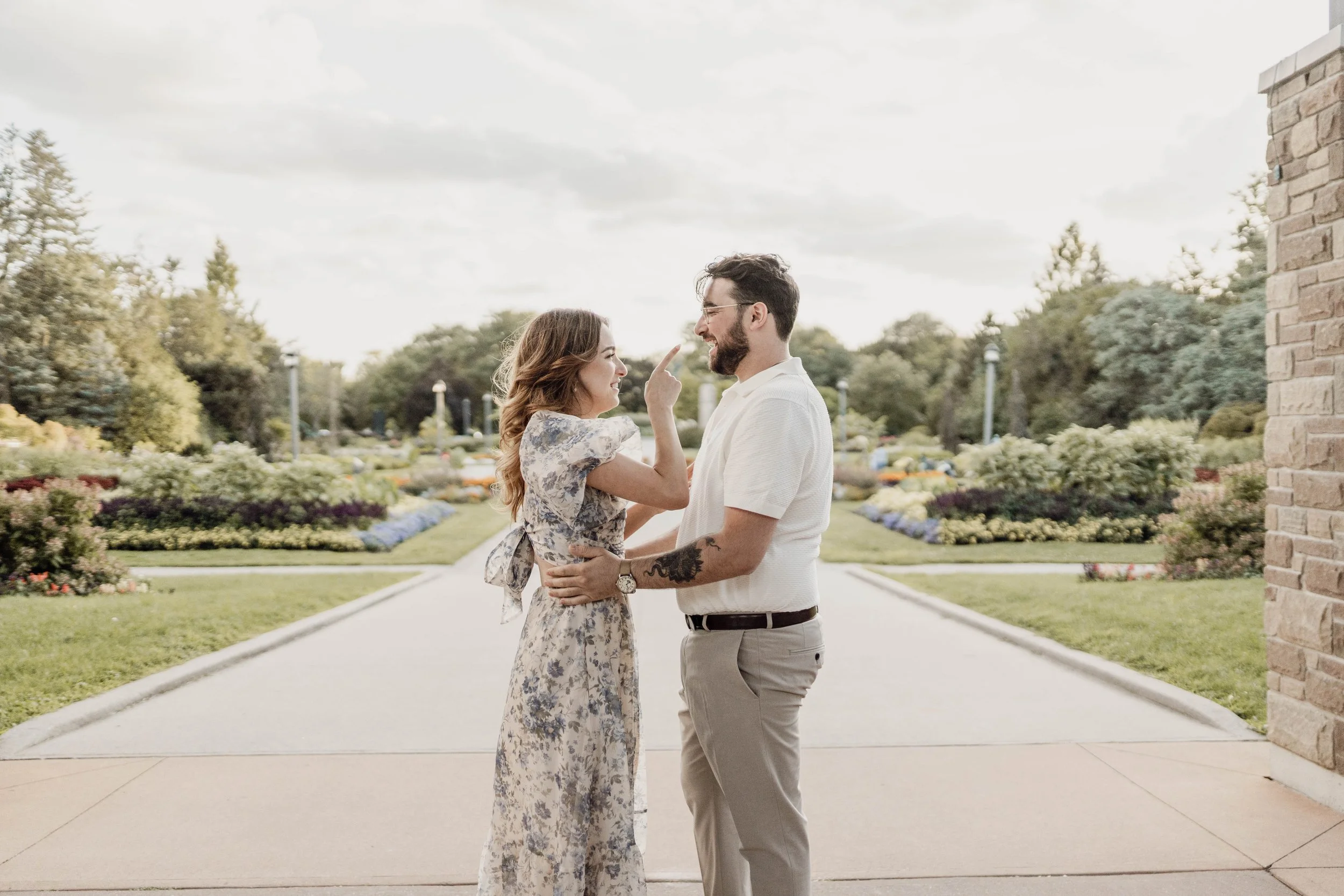 A woman playfully touching the nose of a man in a garden setting with flowers and trees in the background.