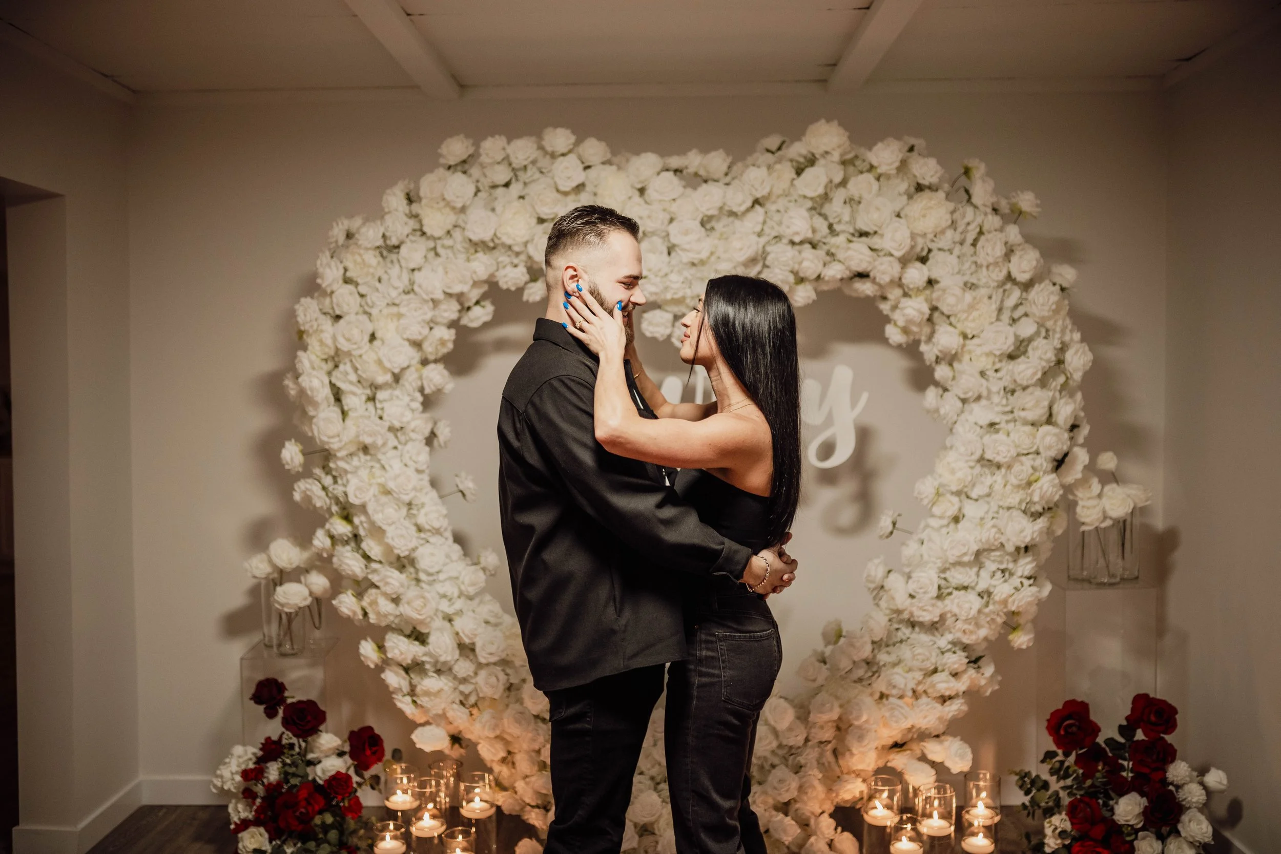 A couple dancing in front of a heart-shaped floral backdrop with candles at the base, surrounded by white and red flowers.