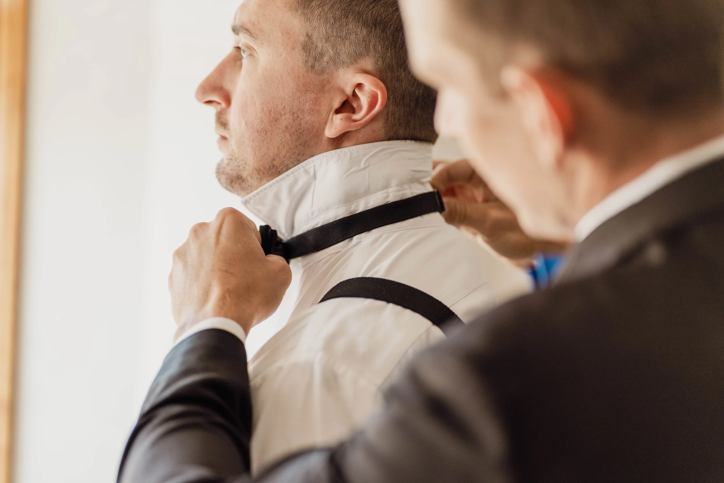 A man helping another man to adjust his black bow tie before a formal event, both wearing suits.