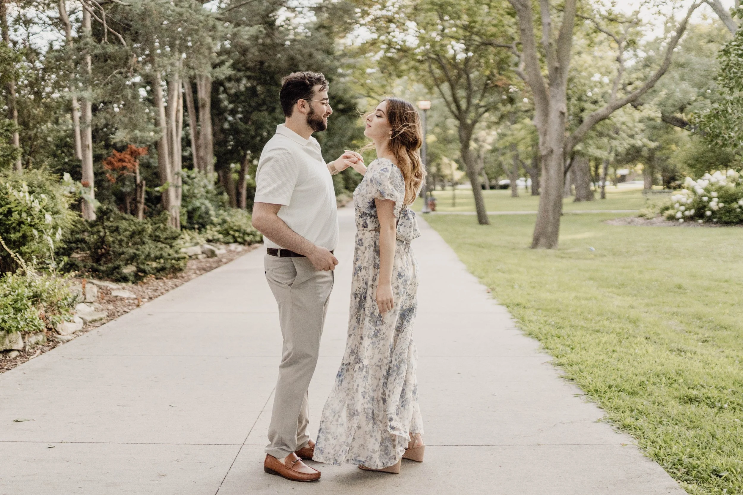A man and woman standing on a park sidewalk holding hands and facing each other, surrounded by trees and greenery.