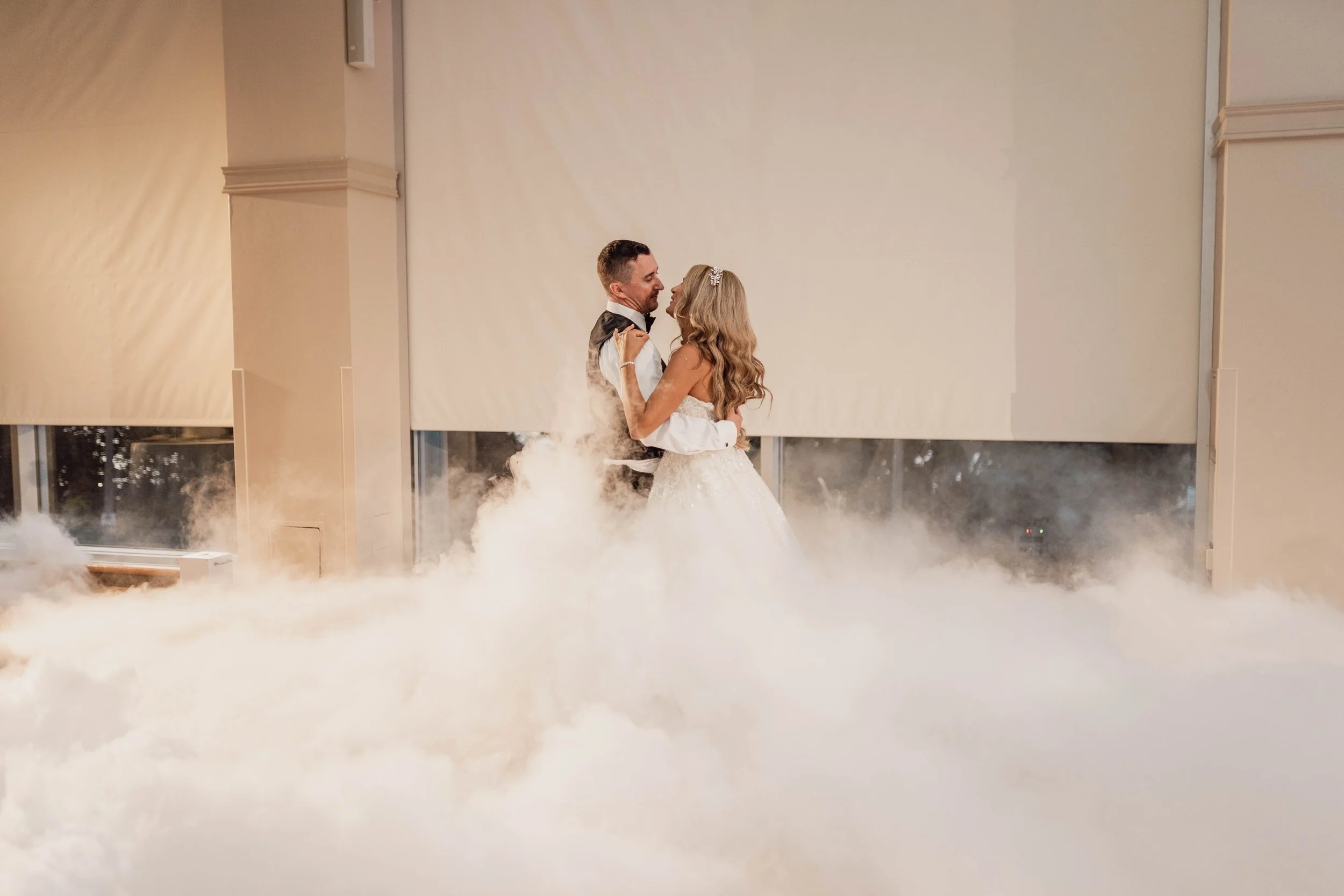 A bride and groom dancing in a dreamy cloud of smoke at their wedding reception, with windows and beige walls in the background.