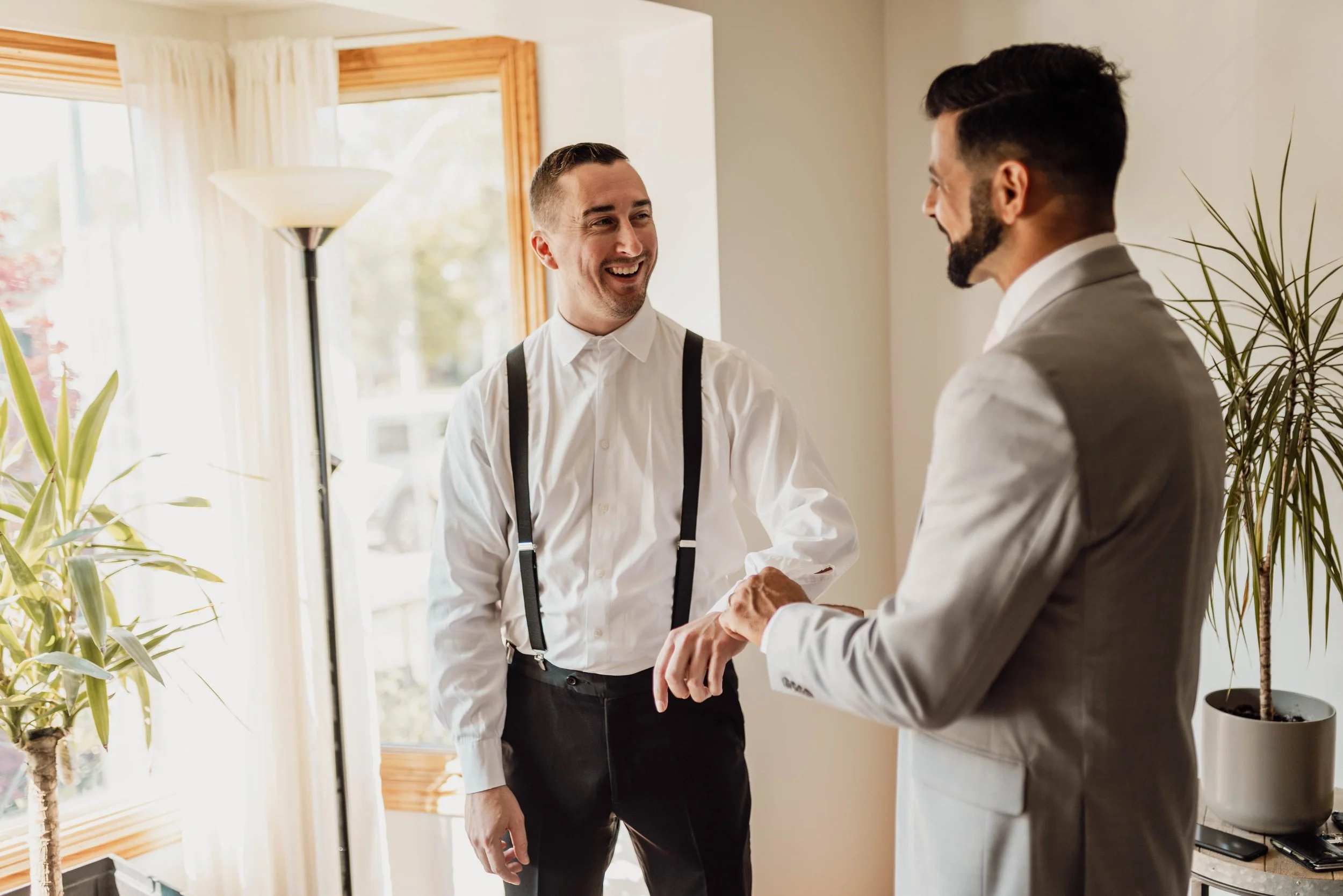 Two men dressed in formal attire are smiling and shaking hands indoors near a window with beige curtains, a potted plant, and natural light.