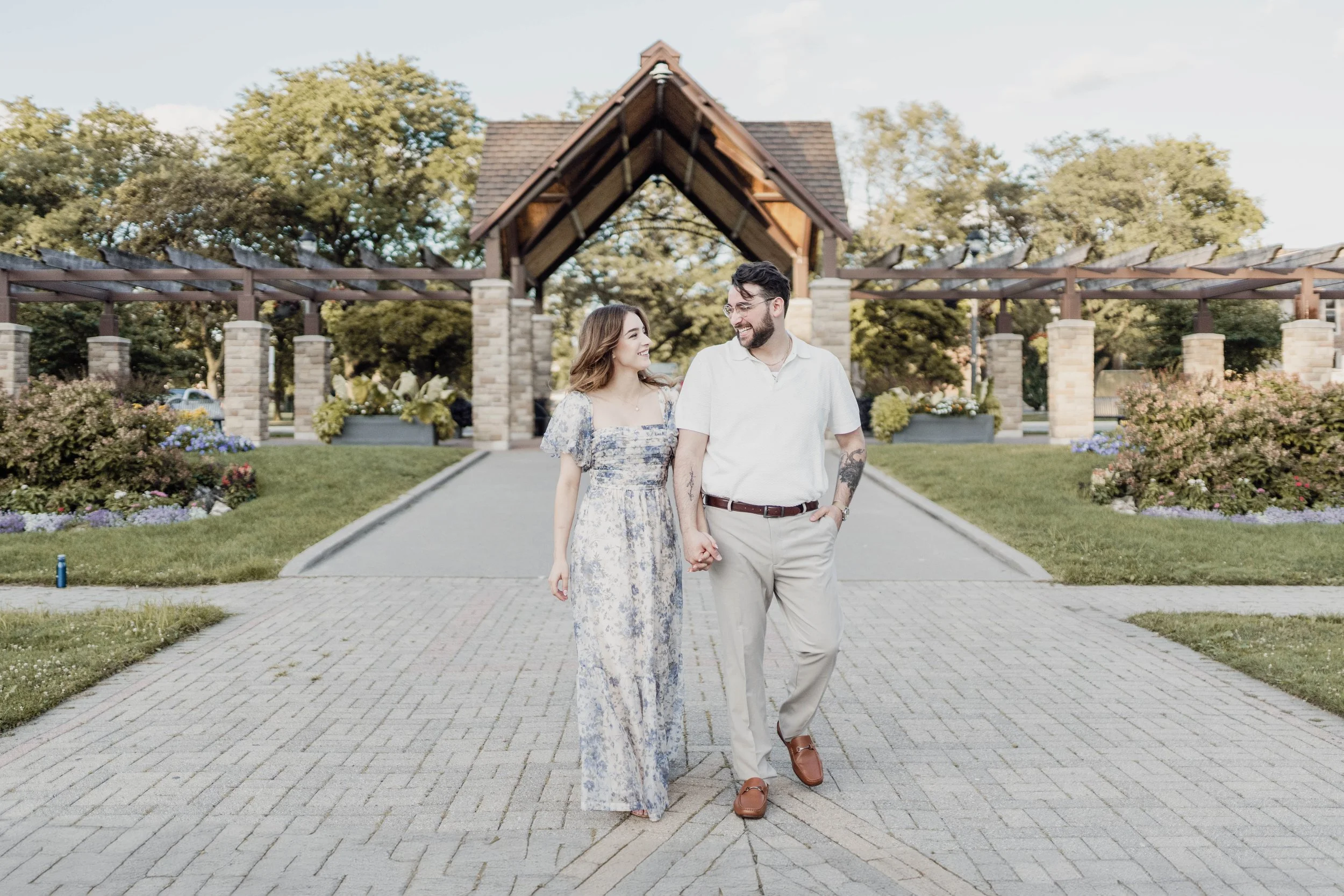 A couple walking hand-in-hand outdoors in a park with trees, flowers, and a stone pavilion in the background.