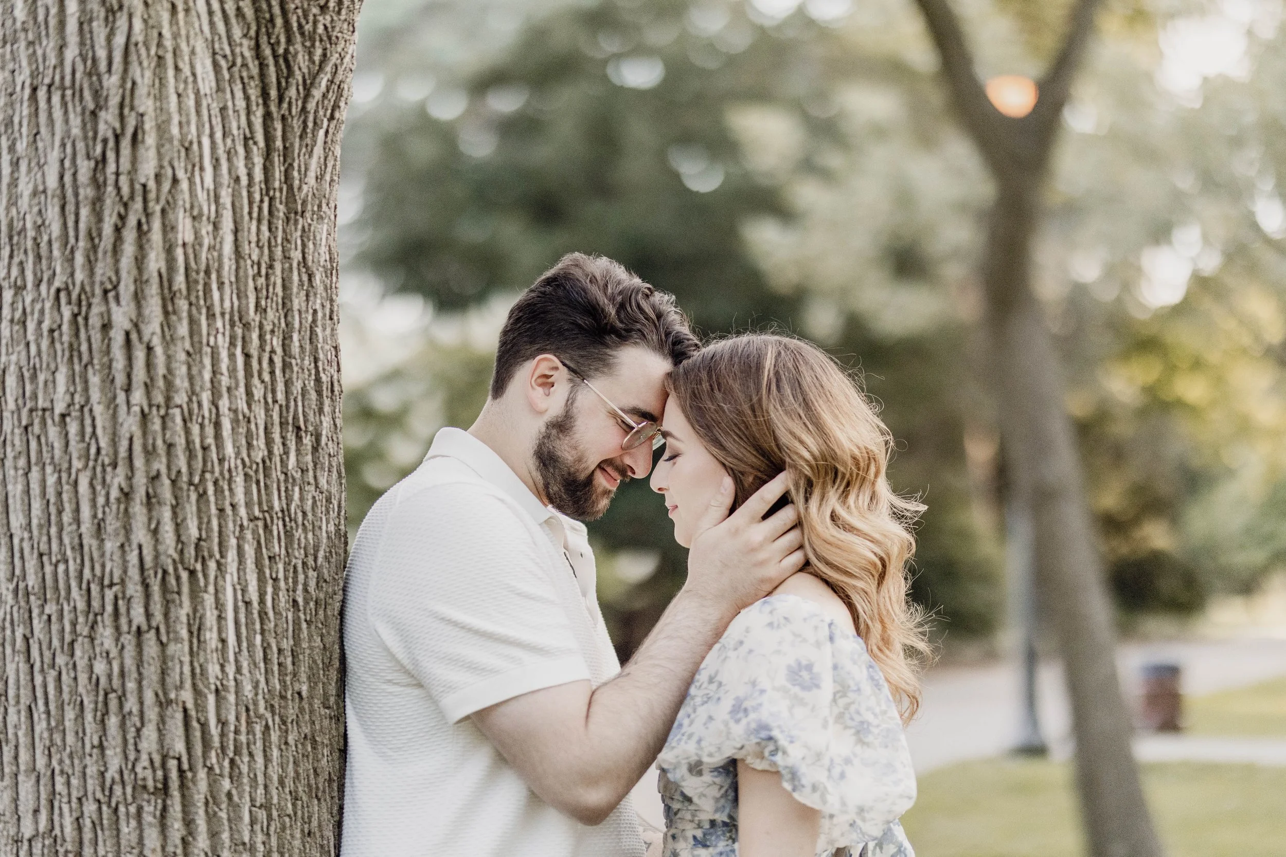 A man and woman close together outdoors, touching foreheads with eyes closed, in front of a tree, with blurred green trees in background.