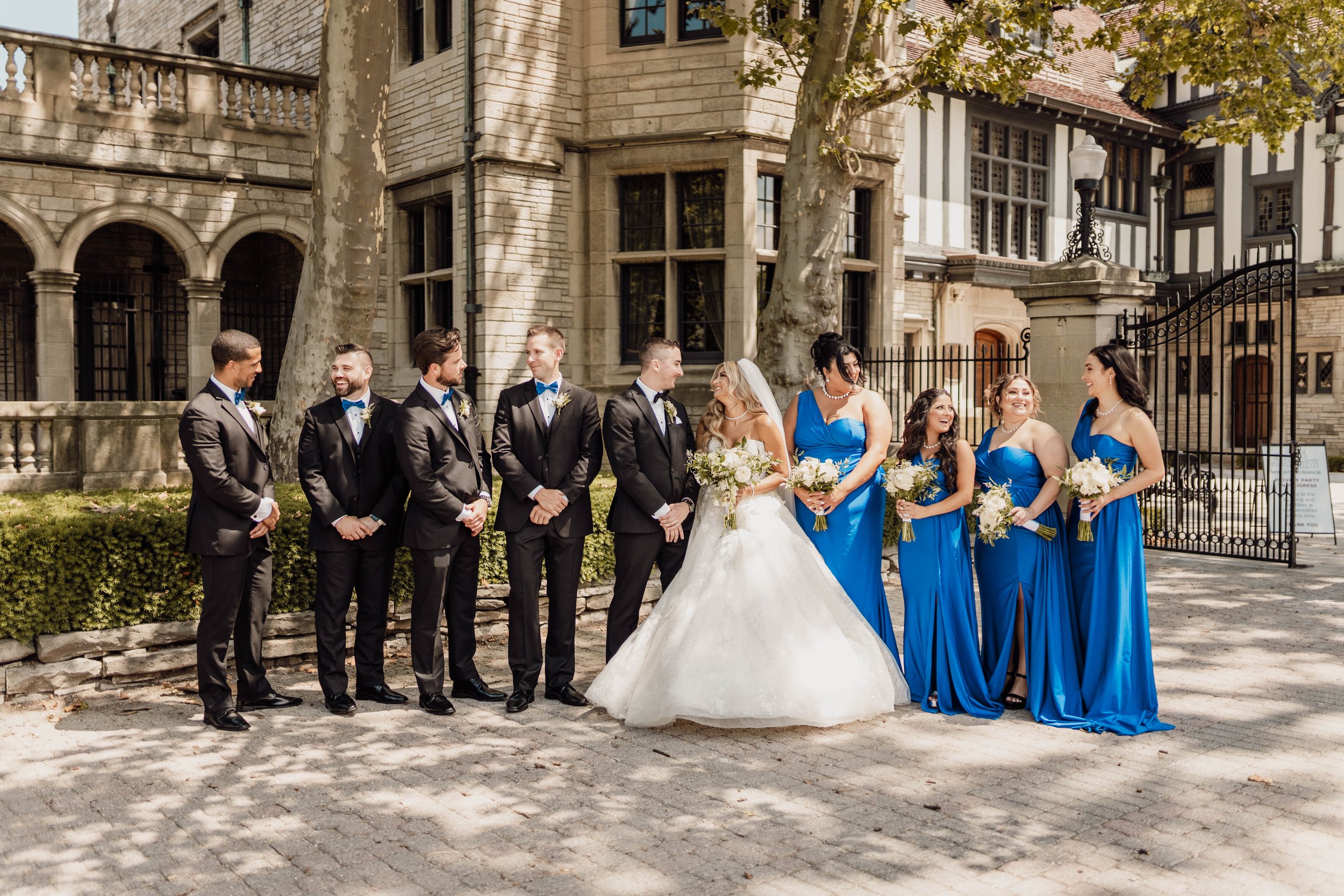 Wedding party standing outdoors near a historic building with trees and shadows. The bride in a white wedding dress holds a bouquet, accompanied by bridesmaids in blue dresses each holding a bouquet, and groomsmen in black suits and bow ties.