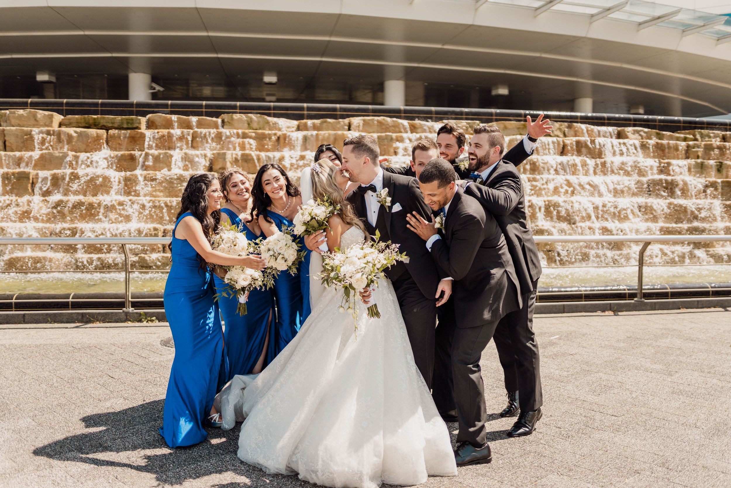 Wedding party of nine, three men and six women, including the bride and groom, standing outdoors in front of a cascading water feature, with the women wearing matching blue dresses and the men in black suits, as they celebrate and share a joyful mome