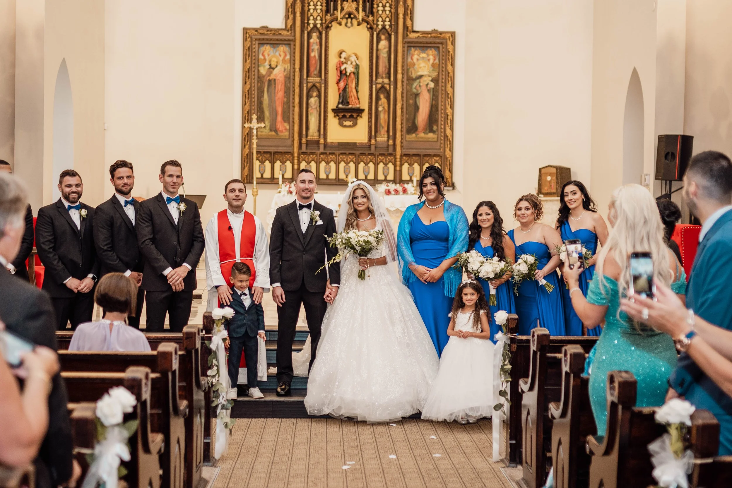 A bride in a white wedding dress standing with guests at a church wedding ceremony, with the wedding party, including bridesmaids in blue dresses and groomsmen in tuxedos, standing behind her.