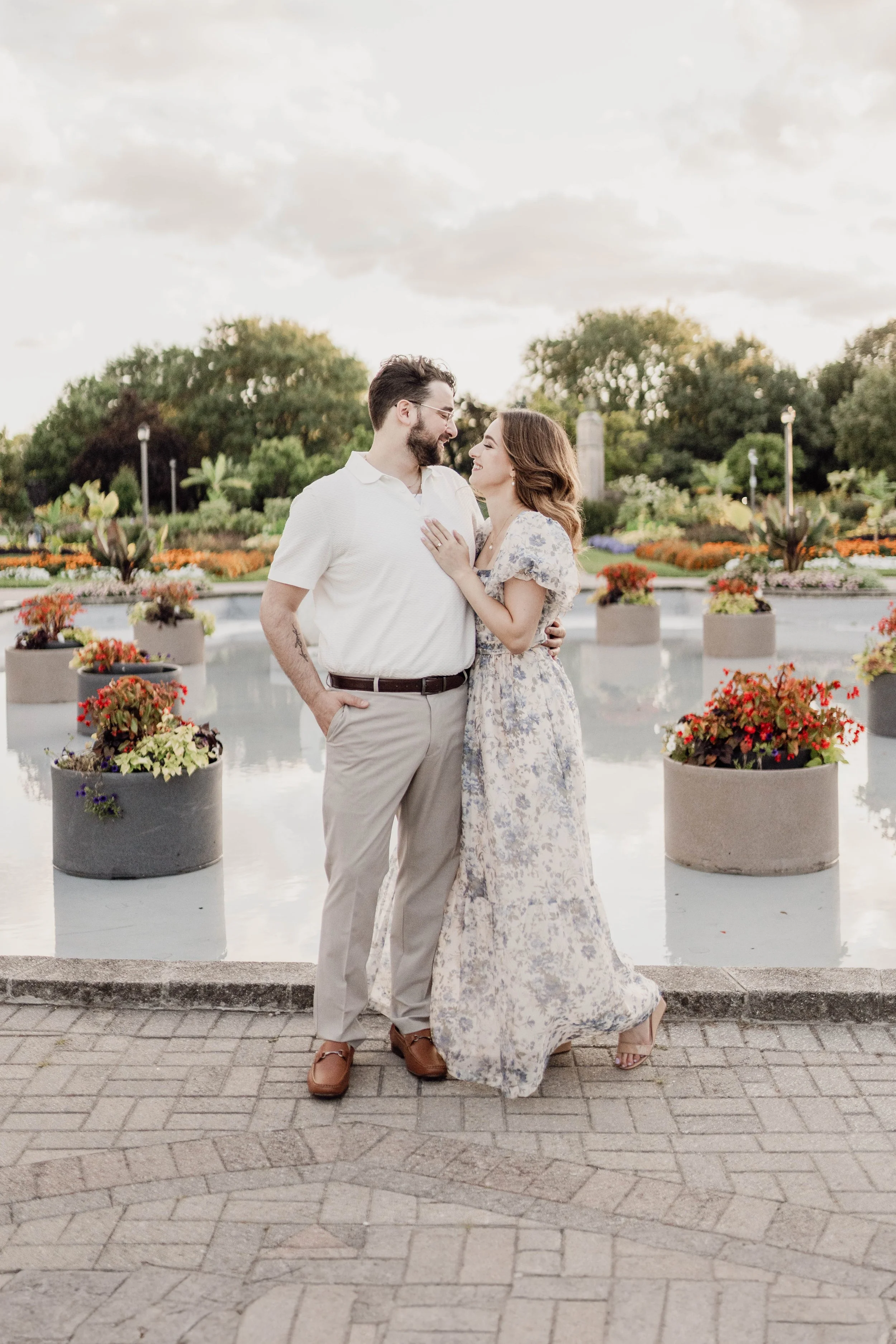 A couple standing by a fountain, gazing at each other, surrounded by potted flowers in a park at sunset.