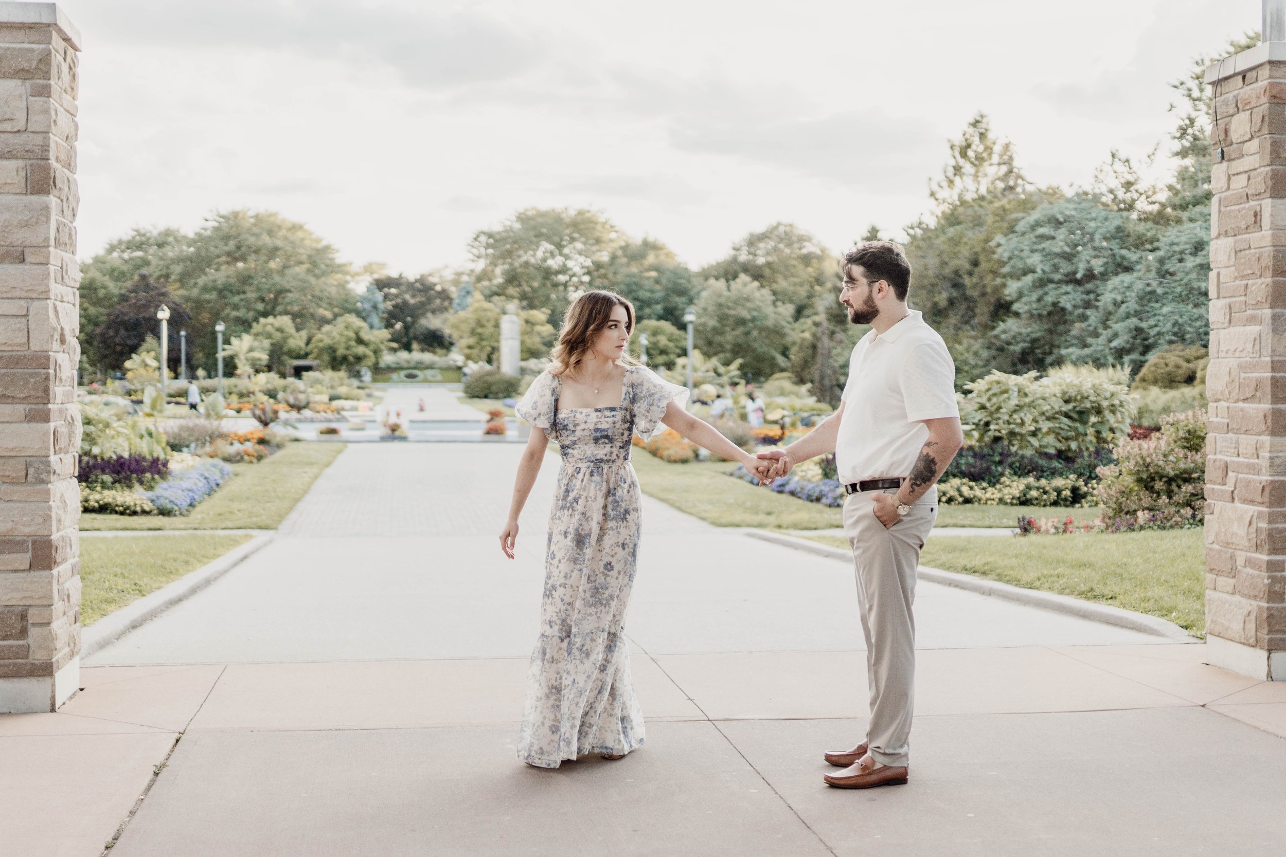A young woman in a floral jumpsuit holding hands with a man in a white shirt and beige pants, standing on a paved walkway in a garden.
