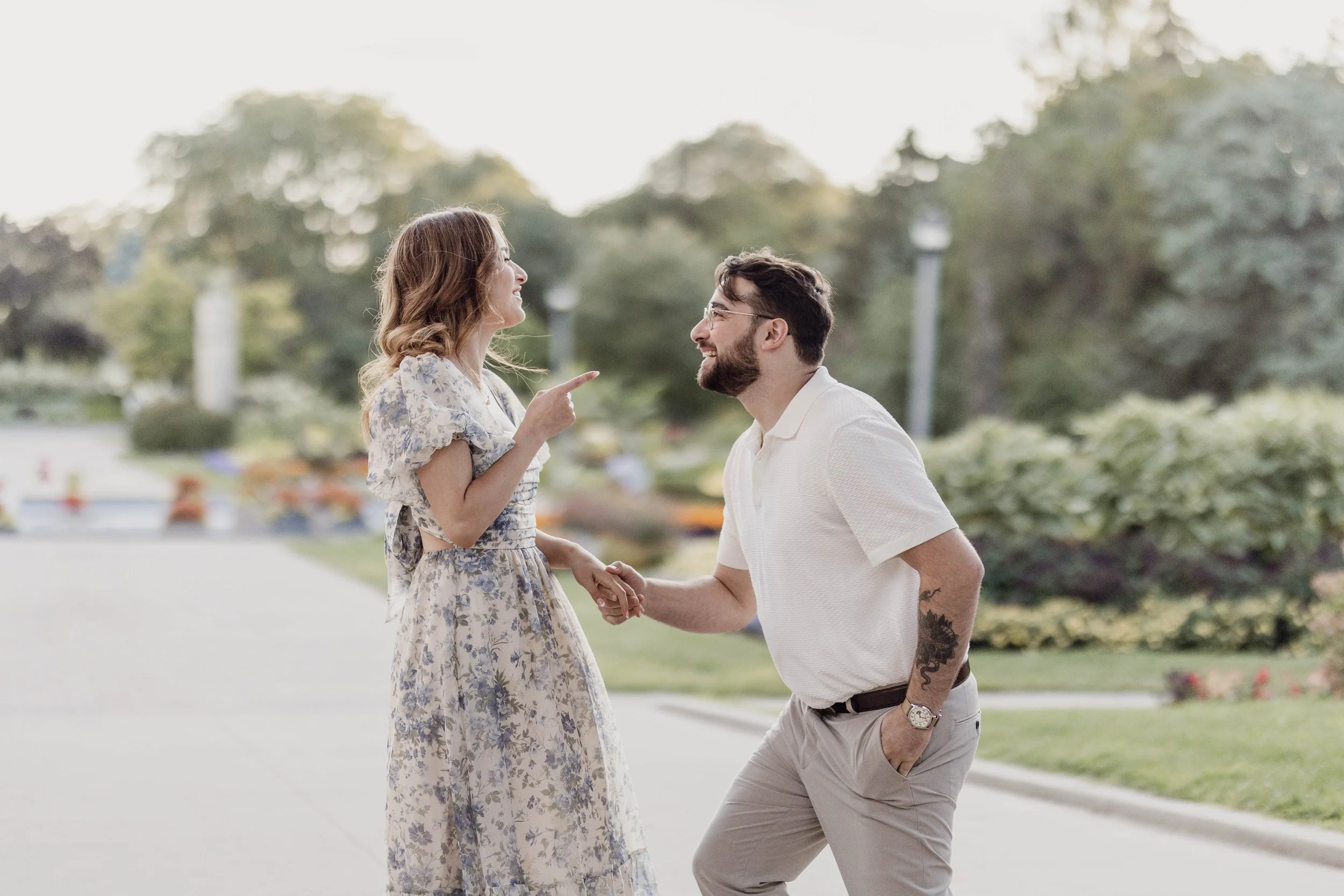A man and woman are smiling and holding hands outdoors in a park, engaging in a playful, affectionate moment.